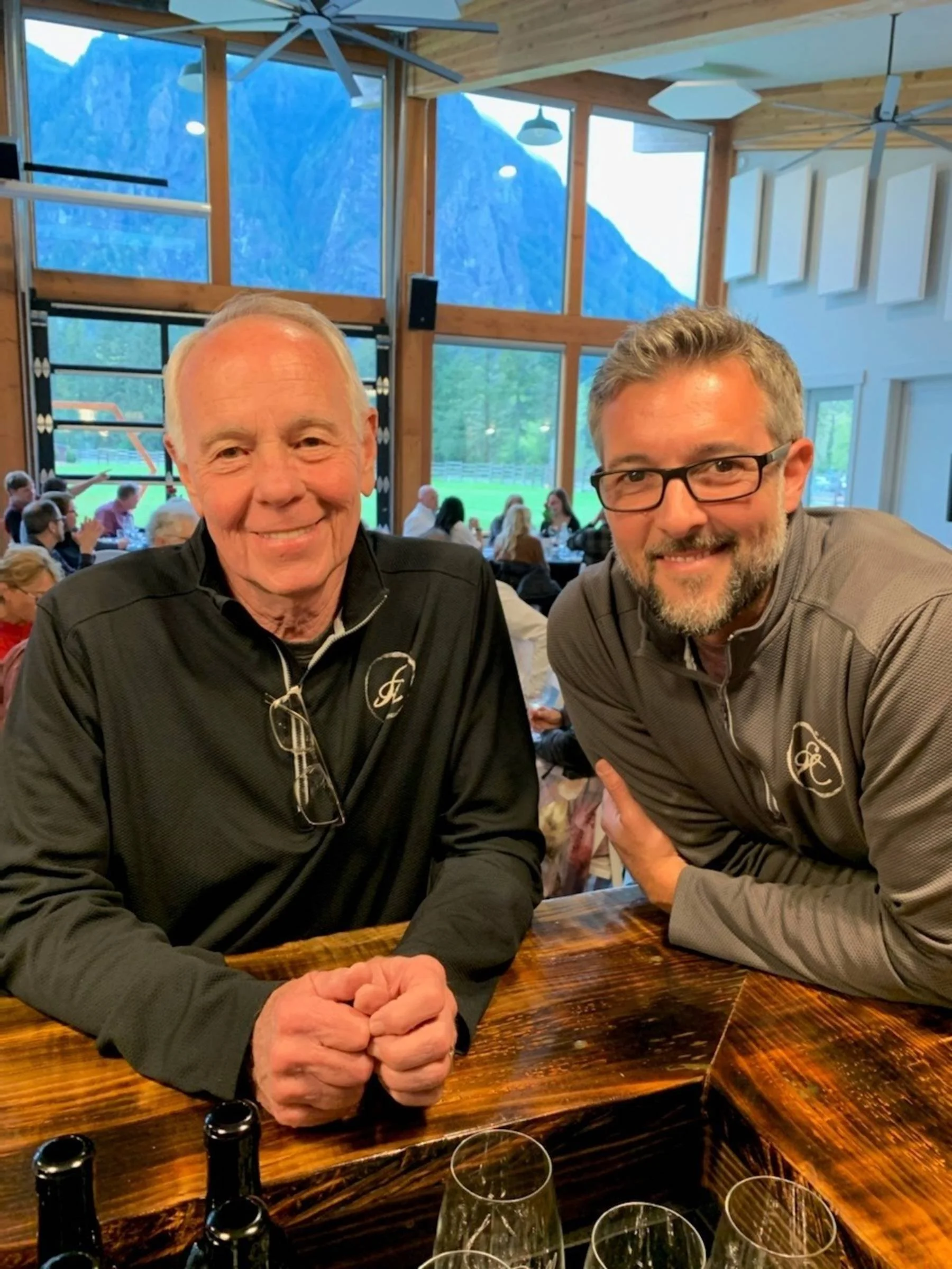Two smiling men posing at a restaurant table indoors, with large windows showing a mountain landscape in the background. One man is older with white hair, wearing a black jacket with glasses hanging from his collar. The other man has gray hair, a beard, glasses, and is wearing a gray jacket. There are bottles and glasses on the wooden table in front of them, and other diners are visible in the background.
