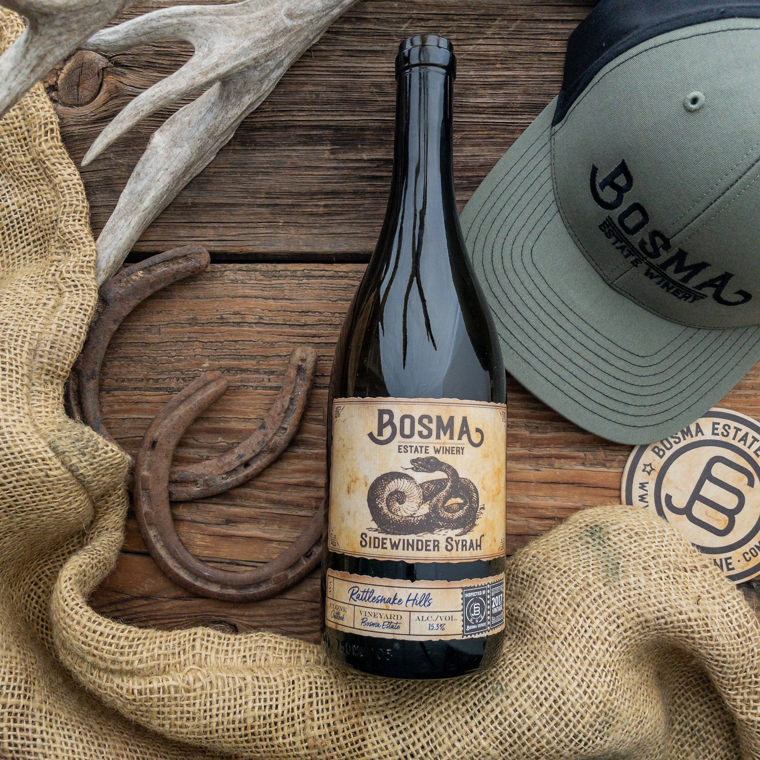 A bottle of Bosma Sidewinder Syrah wine placed on a wooden surface, surrounded by a beige cap, an antler, a piece of burlap fabric, and some rustic chains.