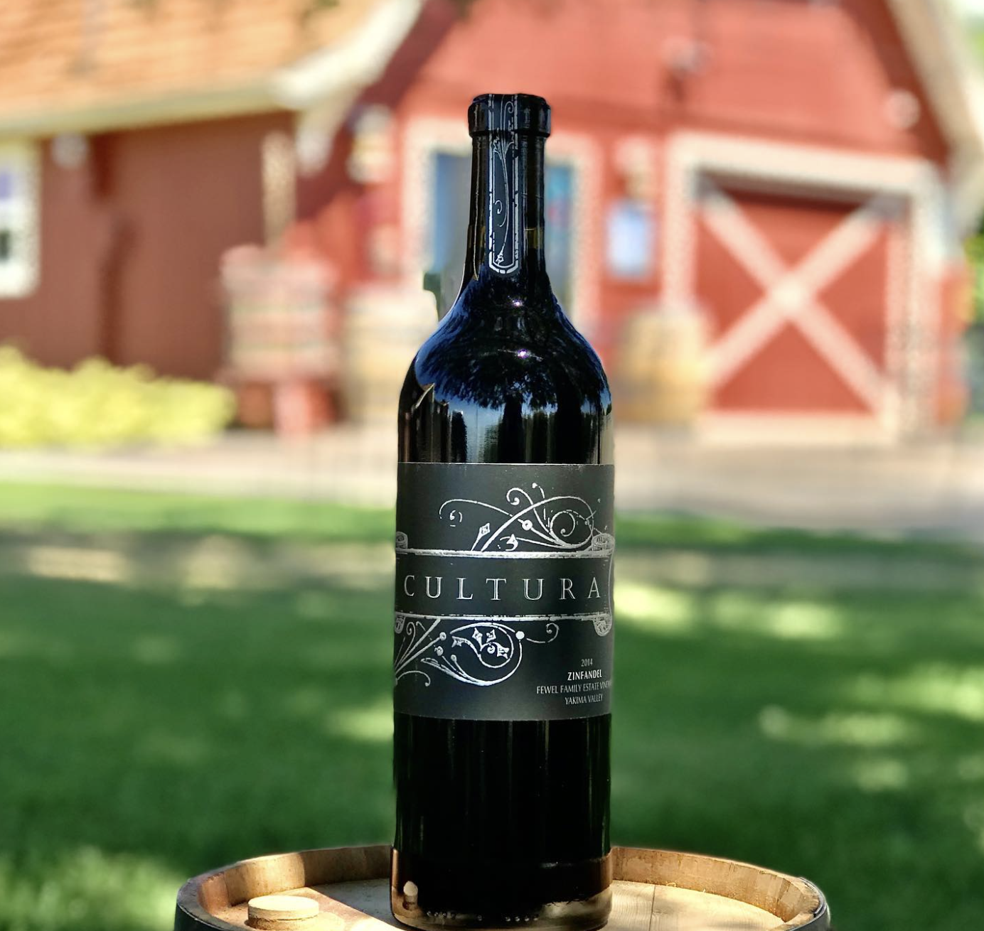 A dark wine bottle labeled 'CULTURA' standing on a wooden barrel outdoors with a red barn and green grass in the background.