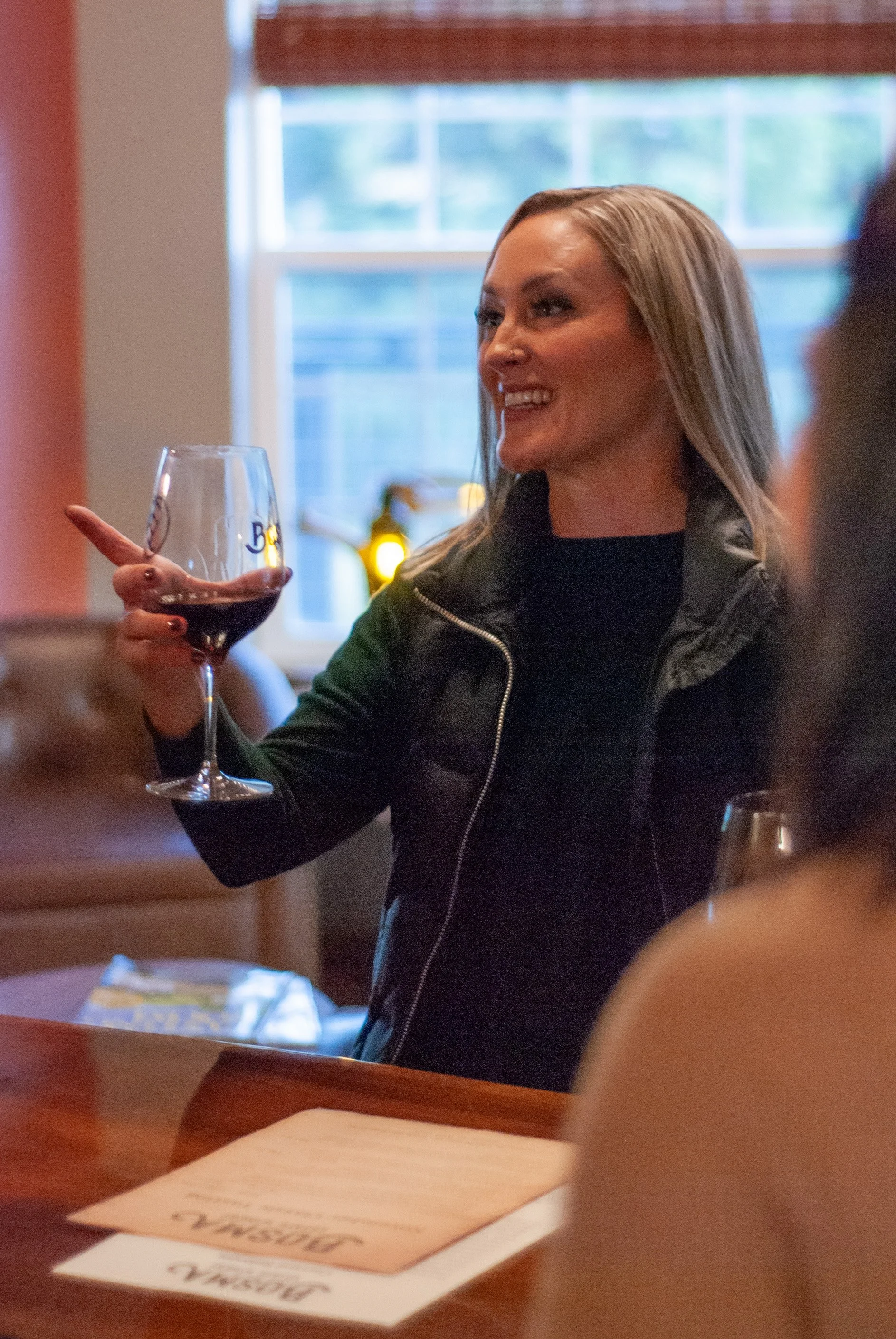 A woman with blonde hair holding a glass of red wine, smiling and engaging in a conversation at a social gathering indoors near a window.