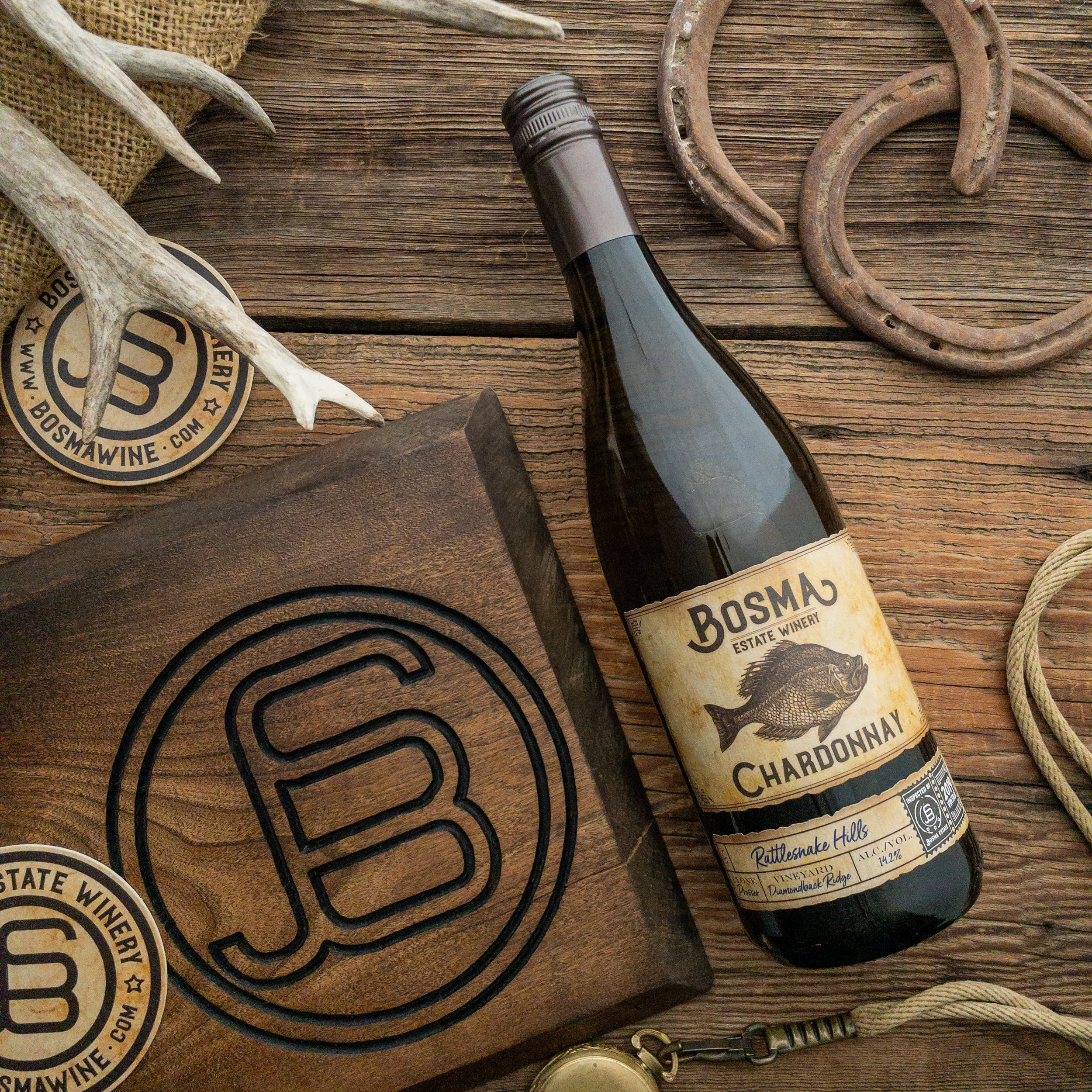 A bottle of Bosma wine lying on a rustic wooden surface surrounded by animal antlers, a horseshoe, a round wire trivet, a round badge, and a locket with a chain.