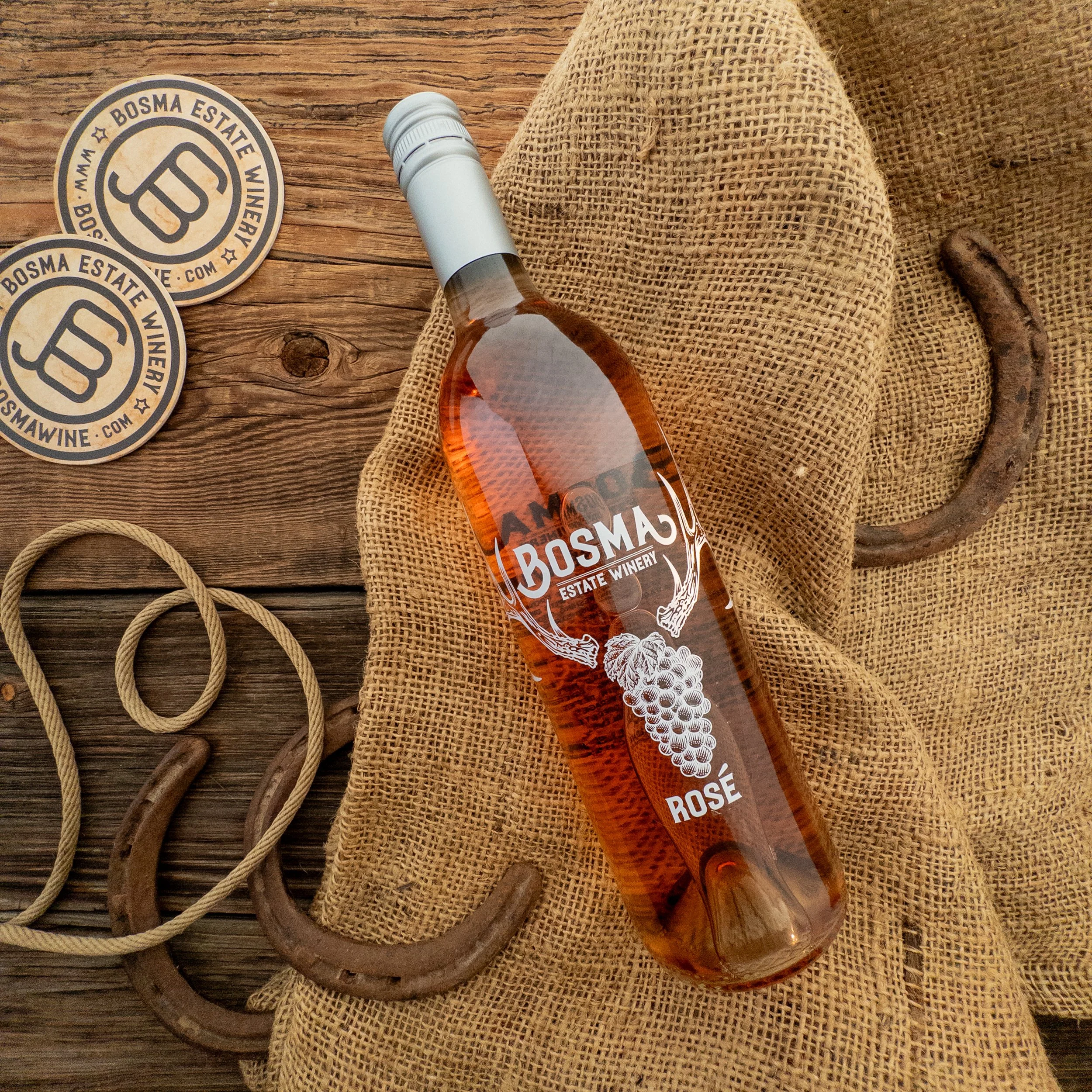 A bottle of Bosma Estate Winery rosé wine on a rustic wooden surface, with pair of burlap fabric and two wine coasters labeled 'Bosma Estate Winery' nearby, along with coiled ropes.