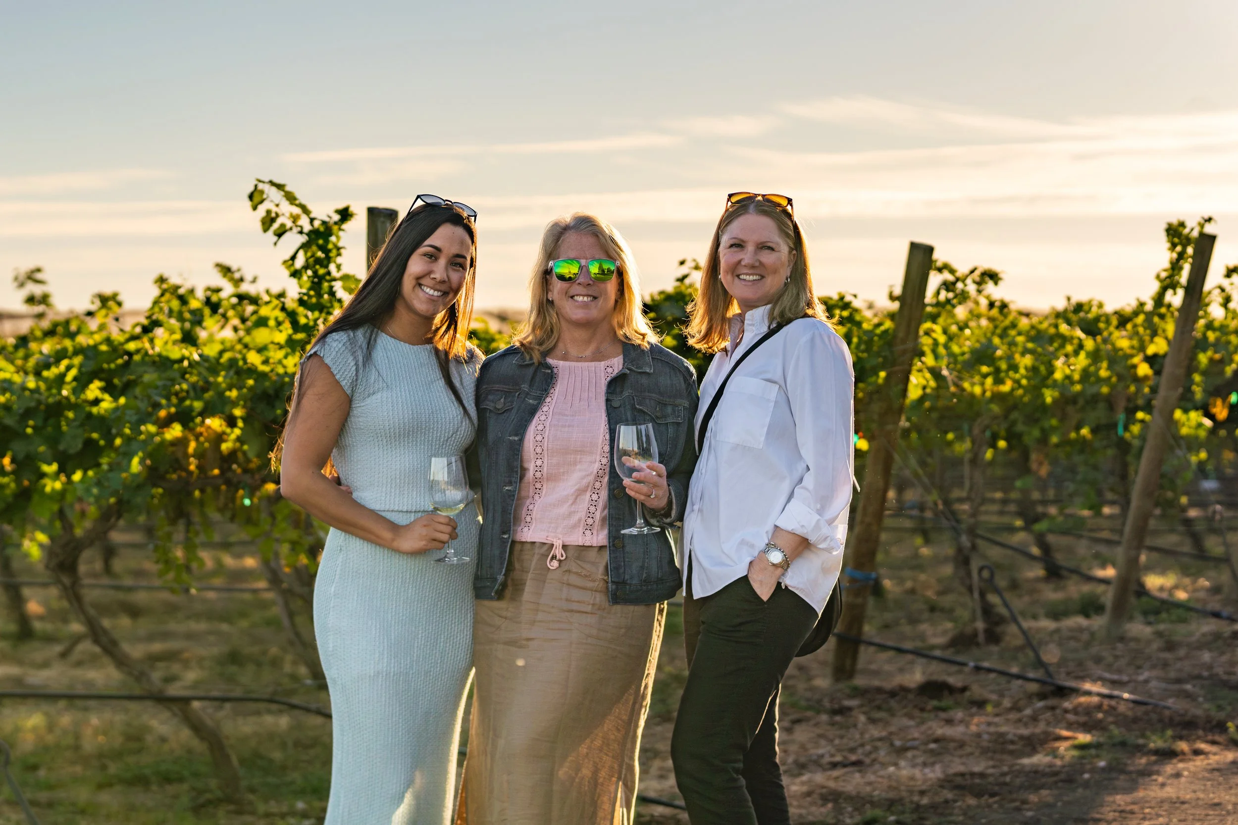 Three women standing together outdoors in a vineyard during sunset, holding wine glasses and smiling.