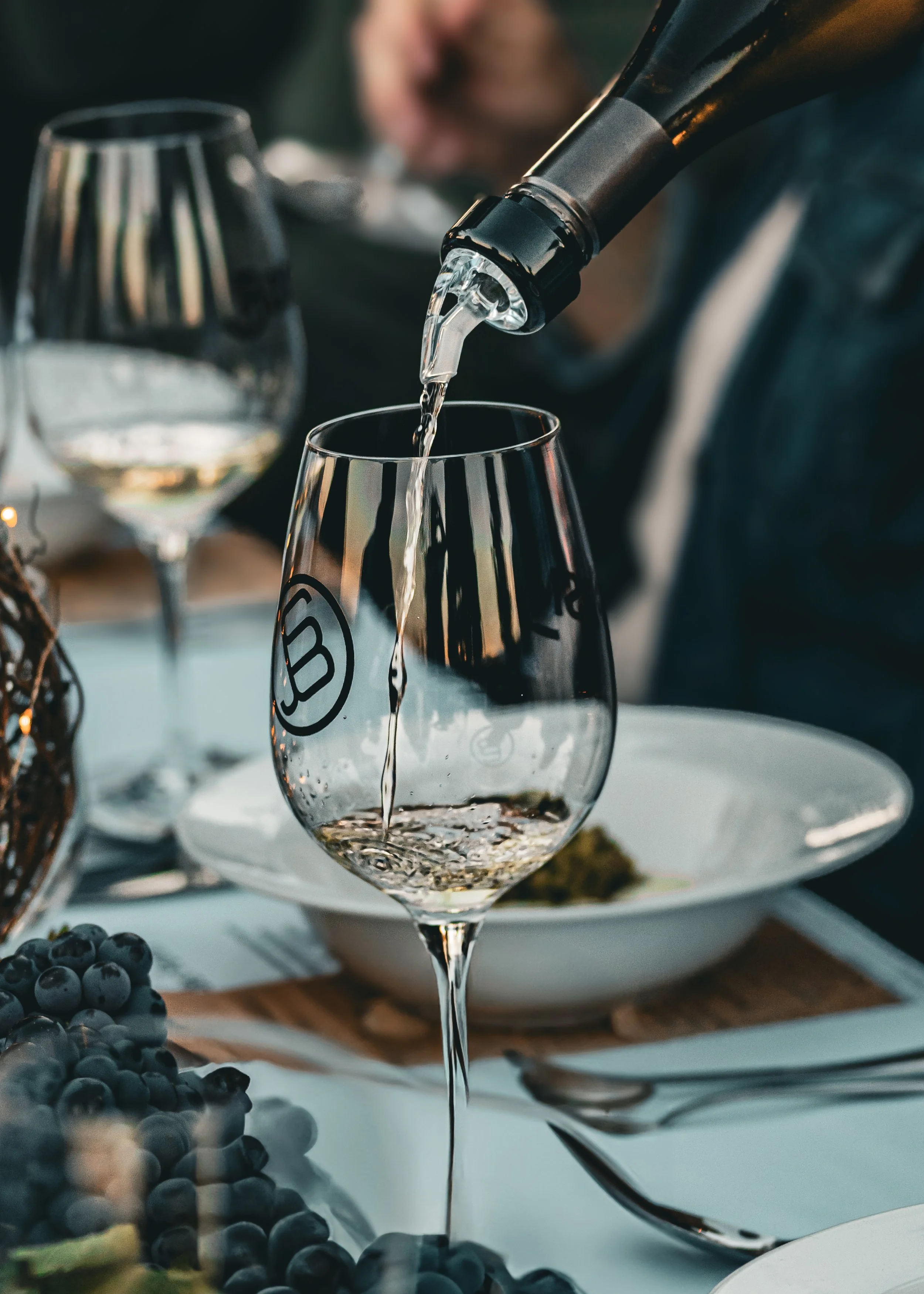 A person is pouring white wine into a wine glass on a table with plates of food and grapes.