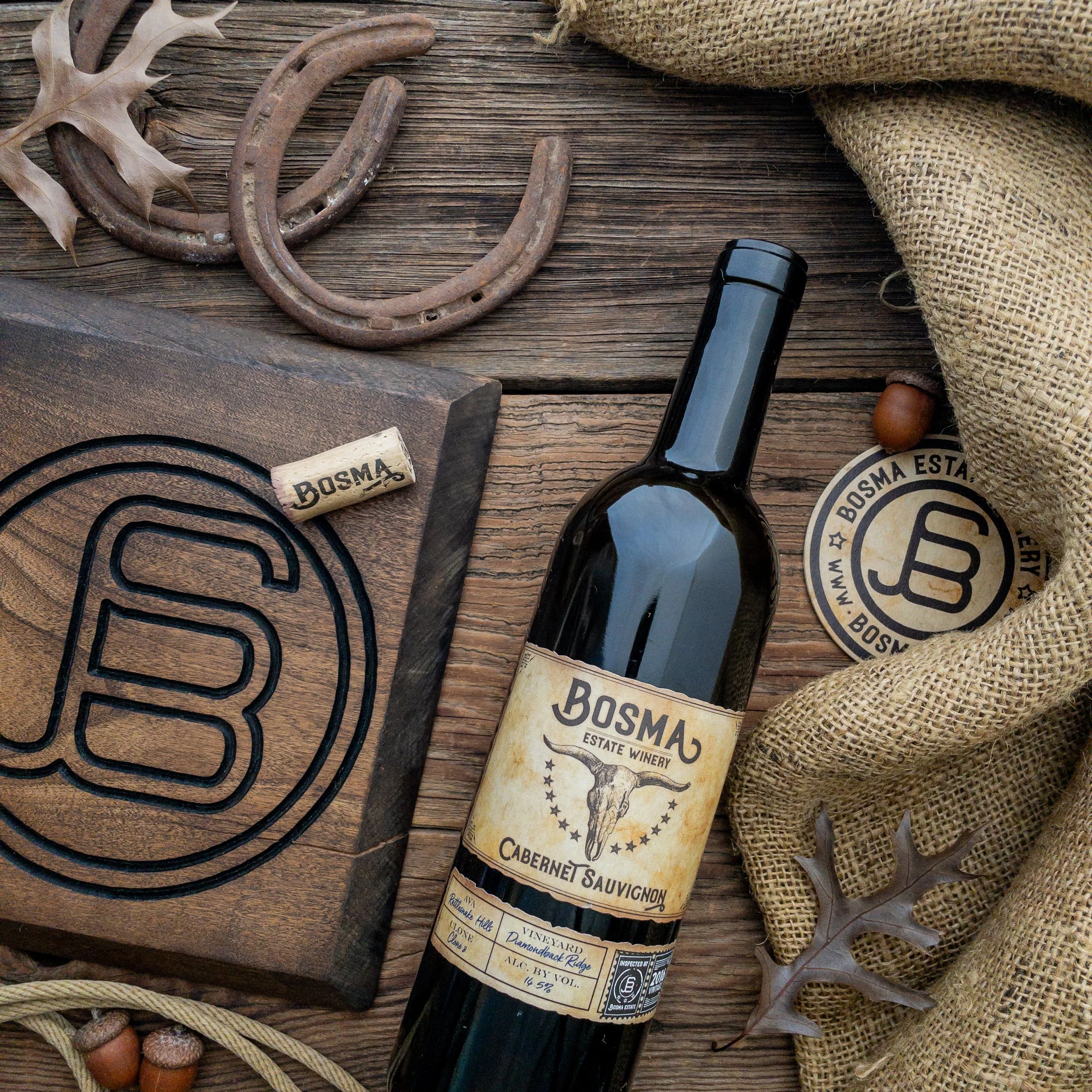 A rustic wooden table with a pair of worn horseshoes, a wine bottle labeled 'Bosma', a cork, a leather coil, a small carved wooden object, some acorns, a round attached label, a burlap bag, and a piece of metal wire.