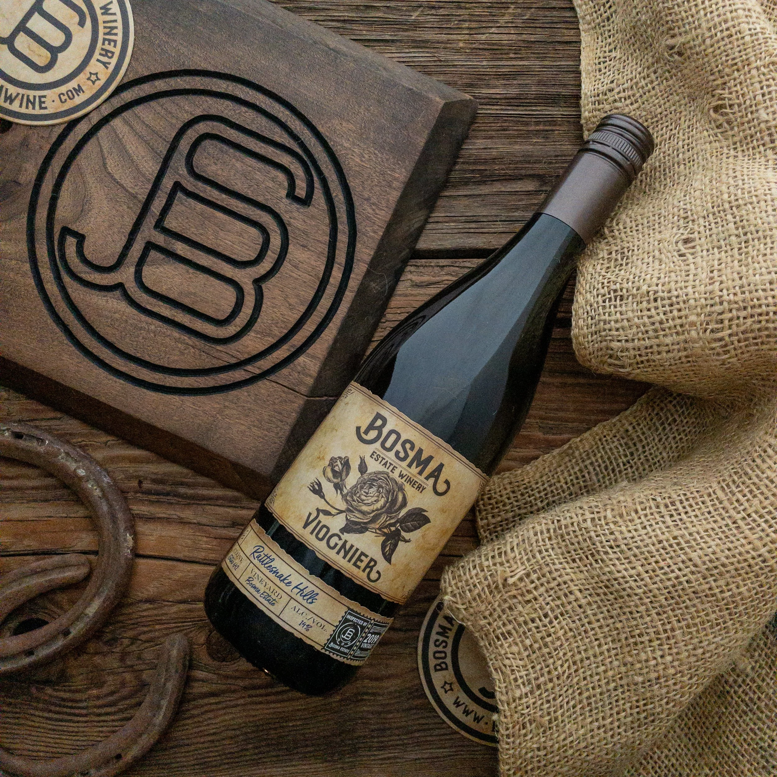 A bottle of Boschma Vineyards Viognier wine lying on a wooden surface, partially covered by a burlap cloth, with a beverage coaster and a metal ring nearby.