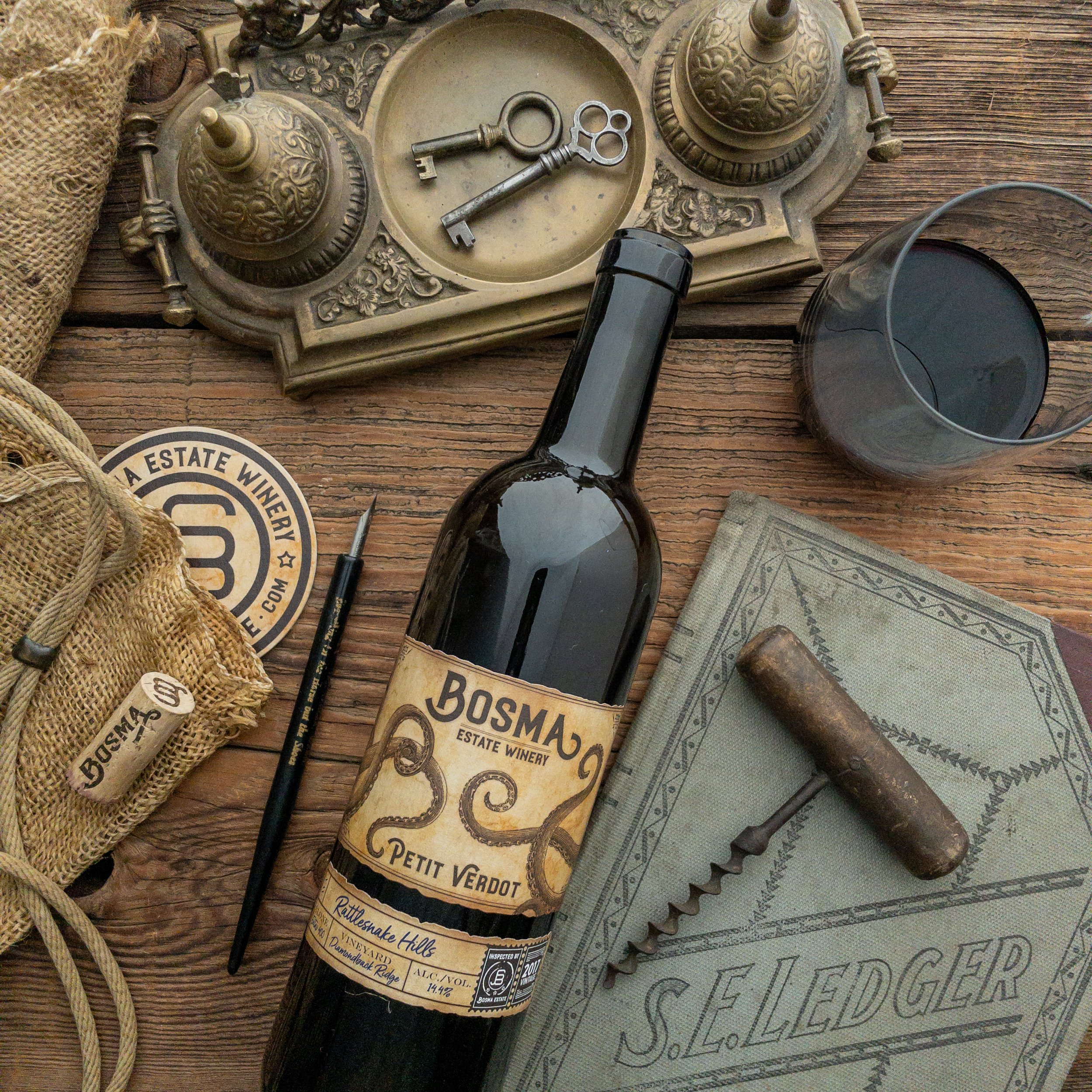 Vintage brass phone, two old keys, a wine bottle, sunglasses, a glass of wine, a cloth, and a wine label on a rustic wooden surface.