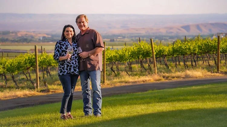 A couple standing on a grassy area in front of a vineyard holding a glass of wine, with rolling hills and a clear sky in the background.