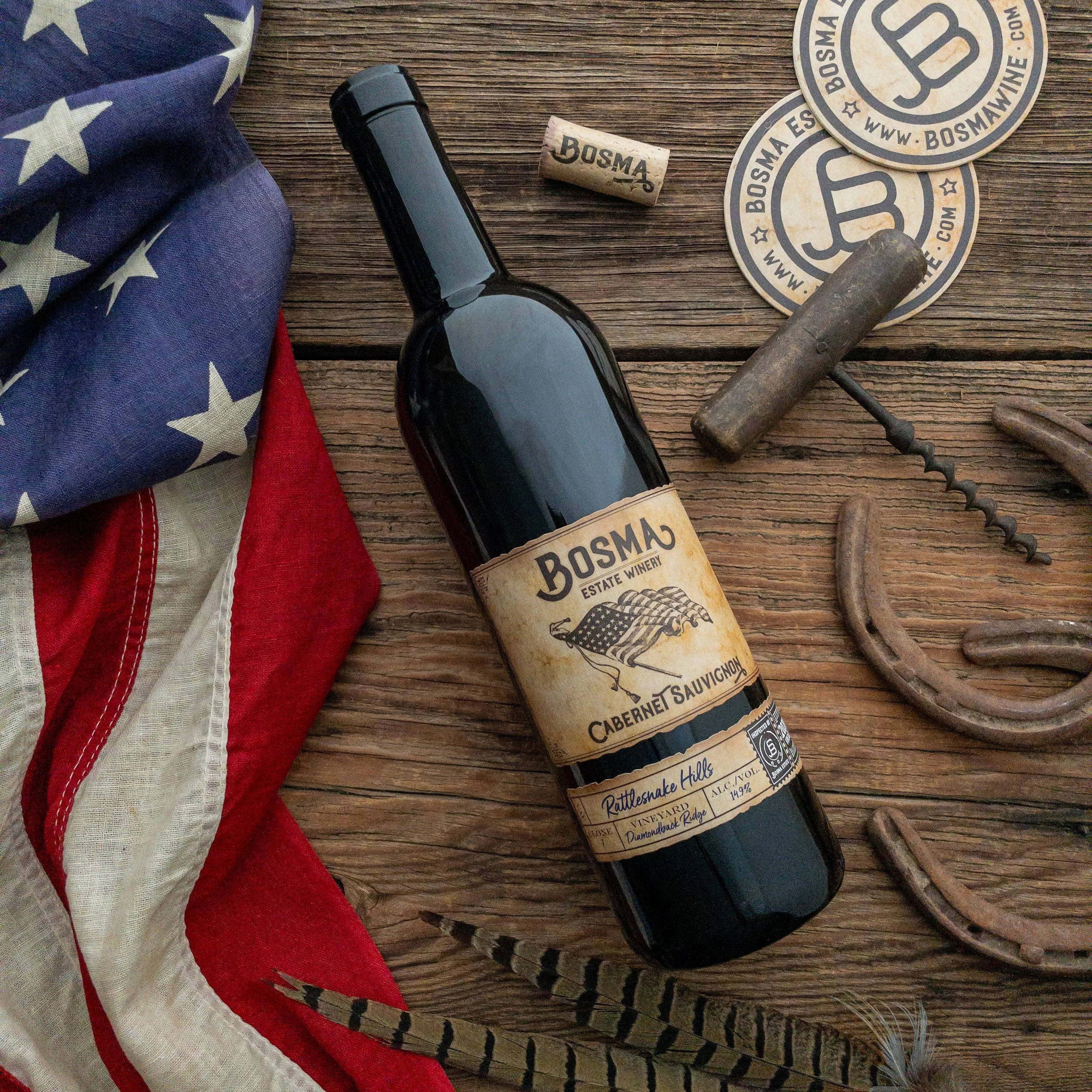 A wine bottle, American flag cloth, wine corks with 'Bosma,' round stickers with 'Bosma Estate Winery,' vintage iron tool, and a rope on a wooden surface.
