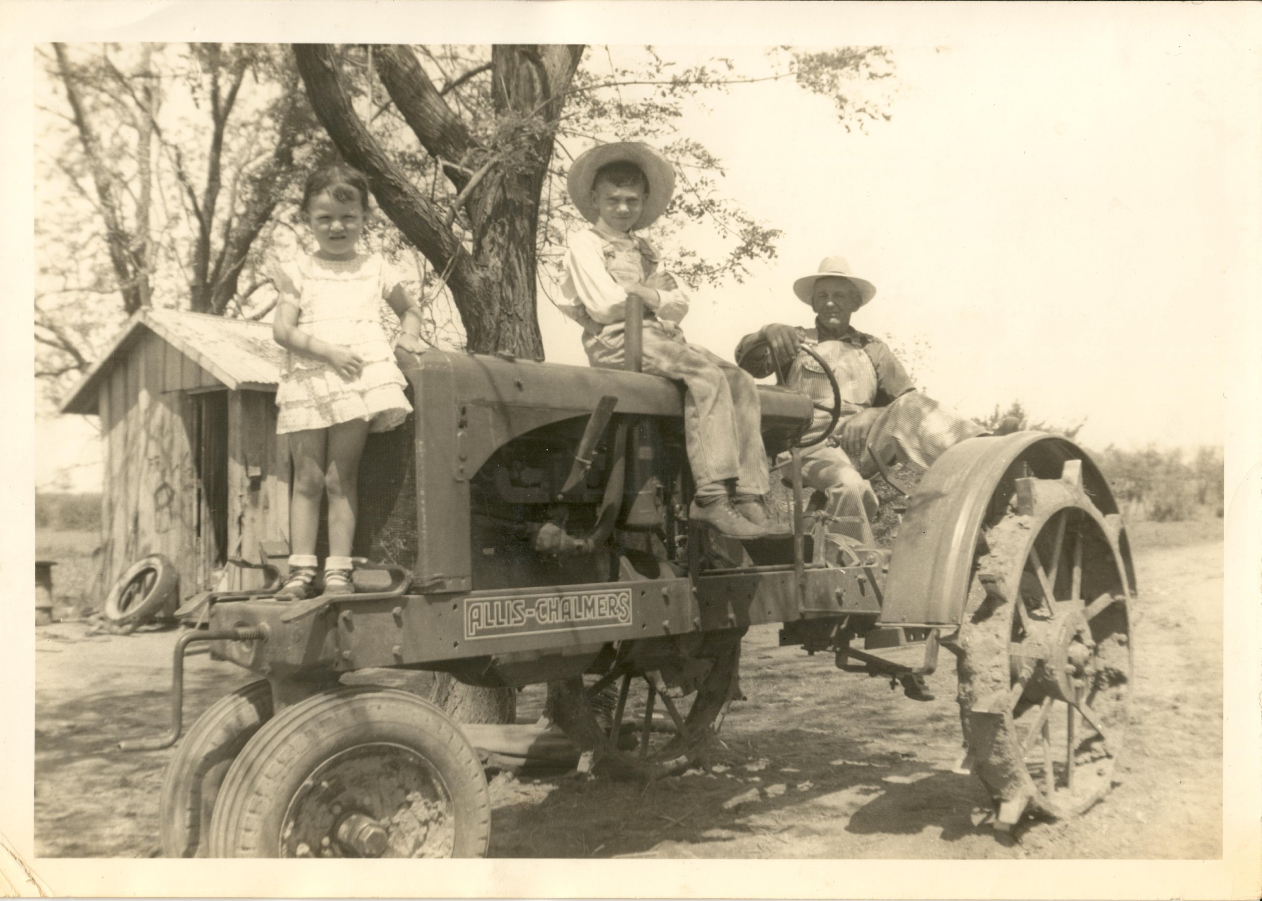 A vintage black-and-white photograph of two children and an older man on a tractor. The children are sitting on the tractor, wearing hats, with the girl on the left and a boy on the right. The man is standing beside the tractor, wearing a hat and a checkered shirt. There are trees in the background, and the tractor has a sign with the text 'ALLIS-CHALMERS'.