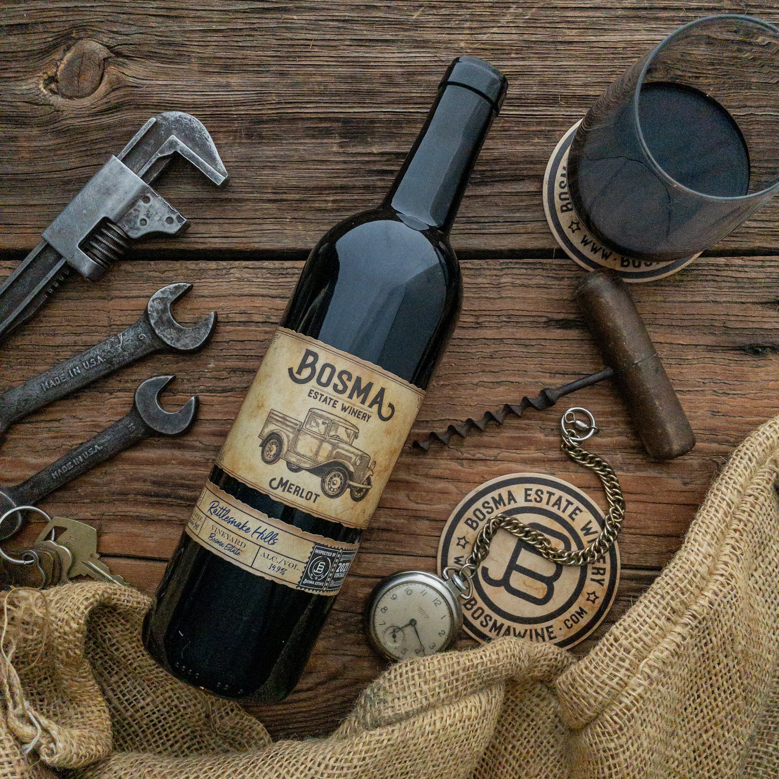 A wine bottle, glasses, and various tools and objects arranged on a wooden surface, including wrenches, a clock, and a sticker or label from Bosma Estate Winery.