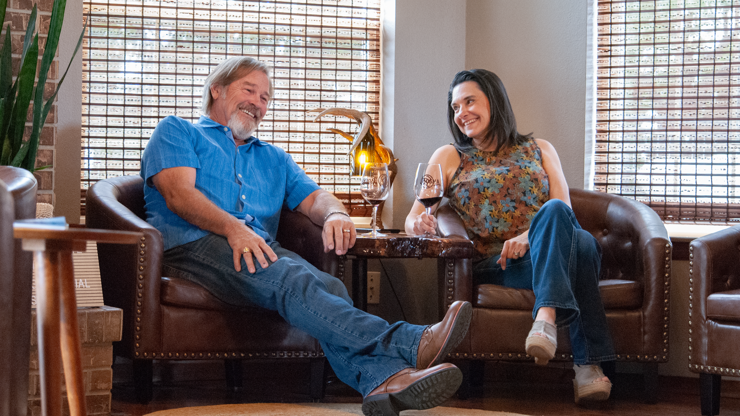 A man and a woman sitting on brown leather armchairs in a cozy room, smiling and holding wine glasses, with bamboo blinds on the windows and a decorative lamp on the table between them.