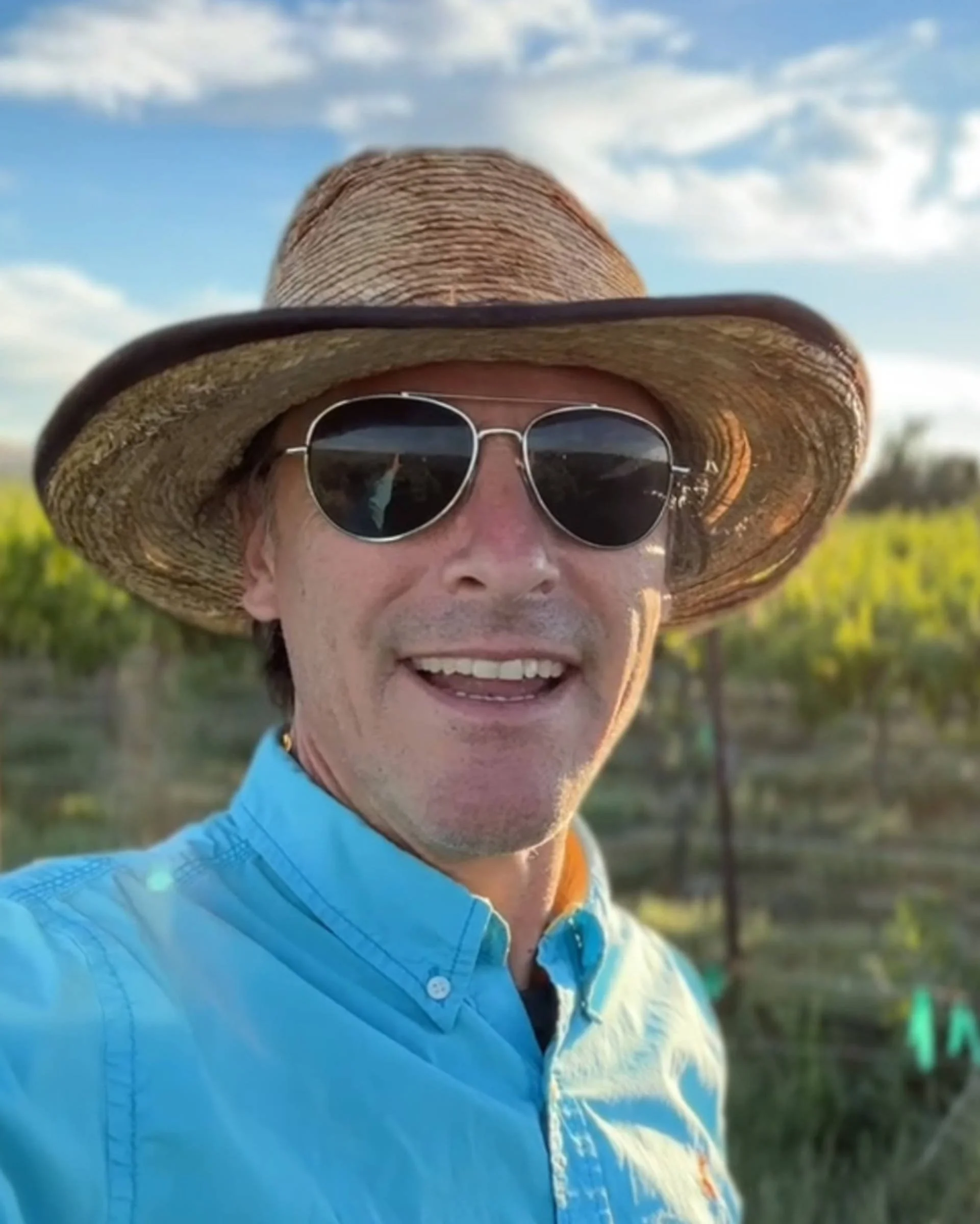 A man is taking a selfie outdoors with a blue sky and white clouds behind him. He is wearing a wide-brimmed straw hat, round sunglasses, a blue button-up shirt, and a gold necklace.
