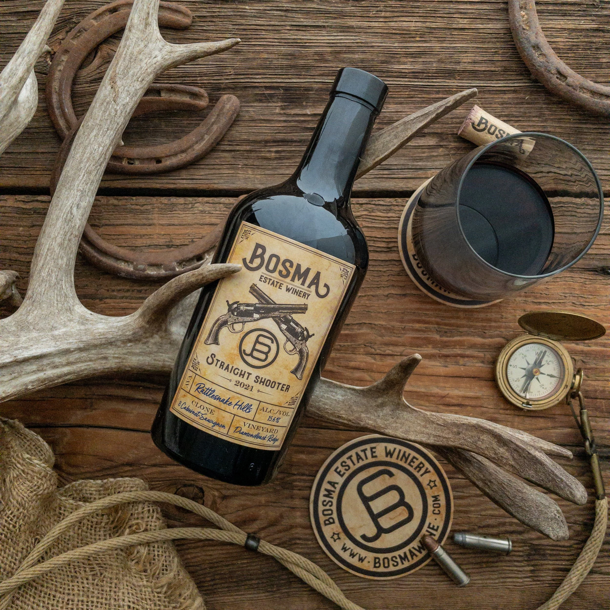 A flat lay of outdoor items including animal antlers, a black wine bottle labeled 'Bosma,' a wine glass filled with red wine, a cork, a pocket watch, and string on a wooden surface.