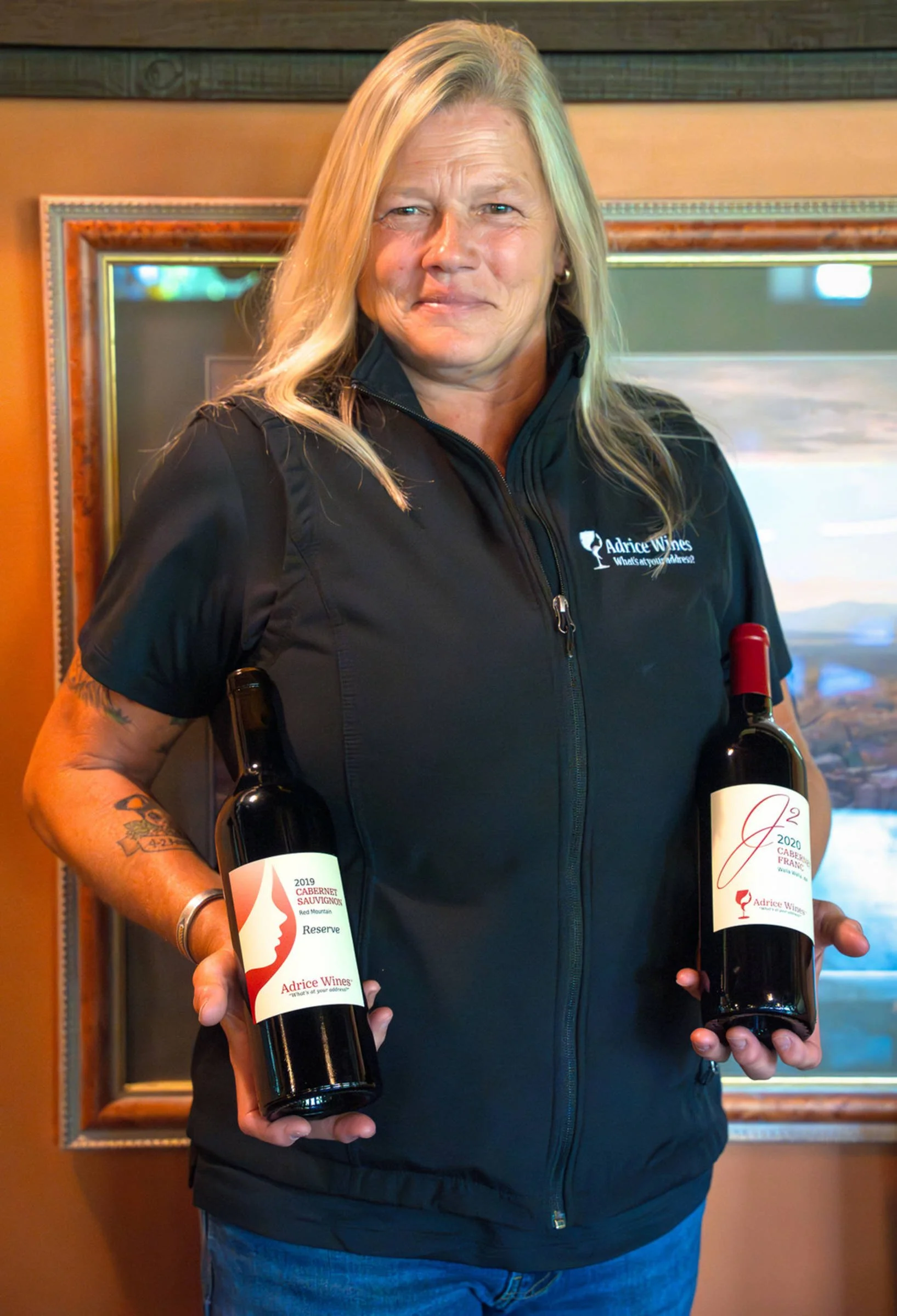 A woman with long blonde hair, wearing a black jacket with the logo and text "Advise Wines," is holding two bottles of wine, one in each hand. The background features a wooden frame and a mirror or window.