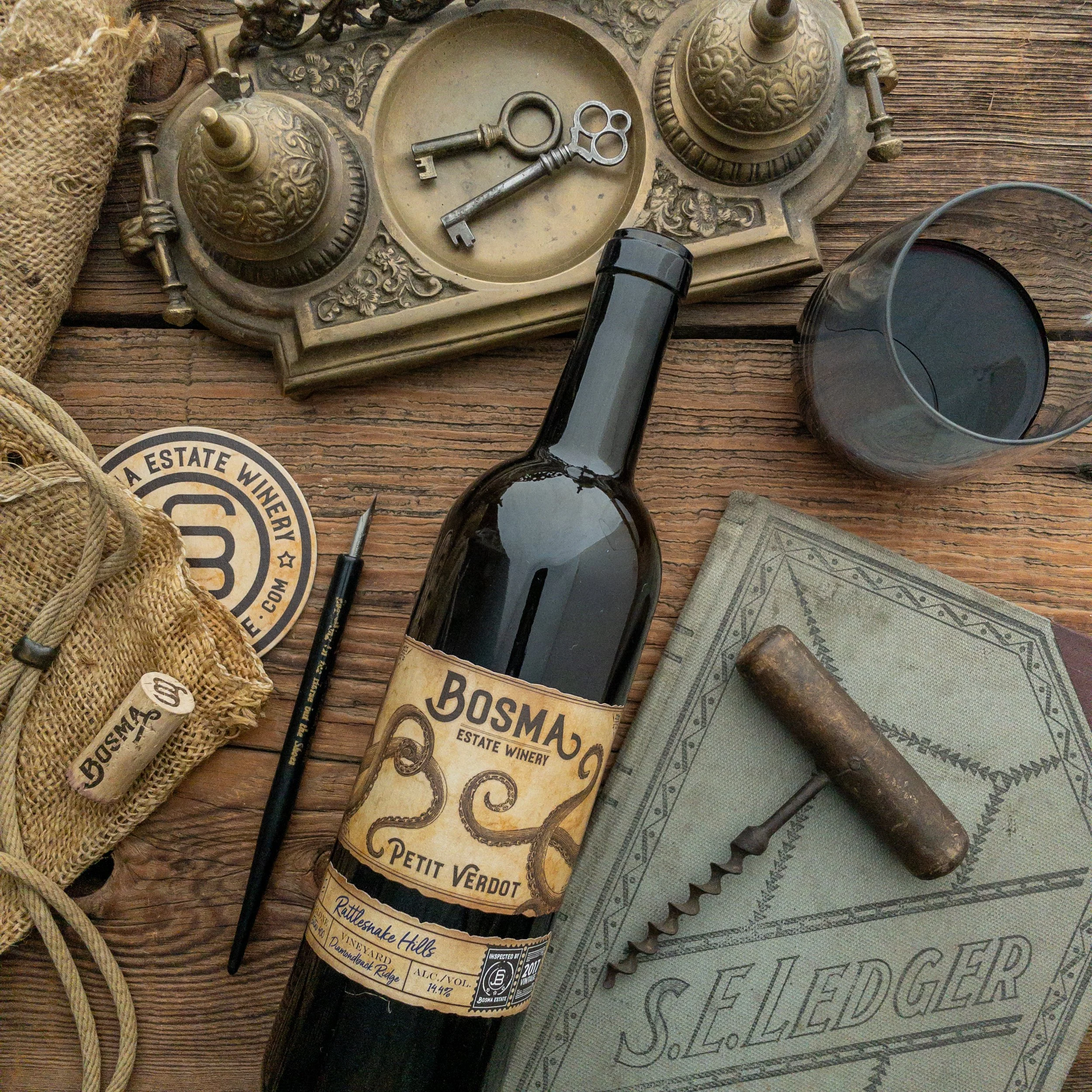 An antique rotary phone, a wine bottle labeled Bosma Petite Verdot, a wine glass, a corkscrew, a map, vintage tags, and a burlap sack placed on a wooden surface.
