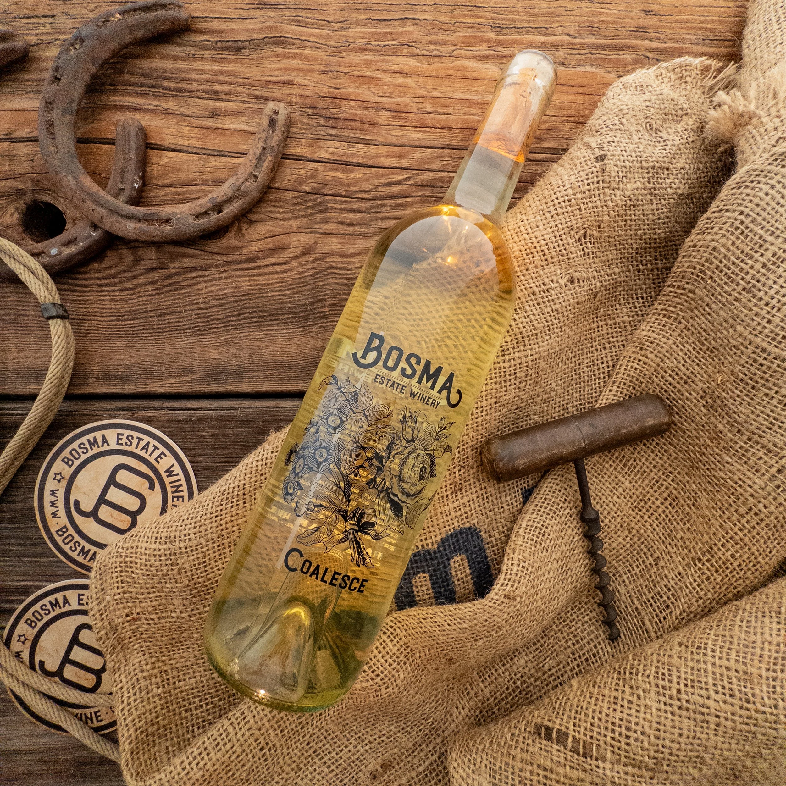 A glass bottle of Bosma Estate Winery Coalesce wine resting on burlap fabric next to an antique metal horseshoe and a round Bosma Estate Winery sticker on a wooden surface.