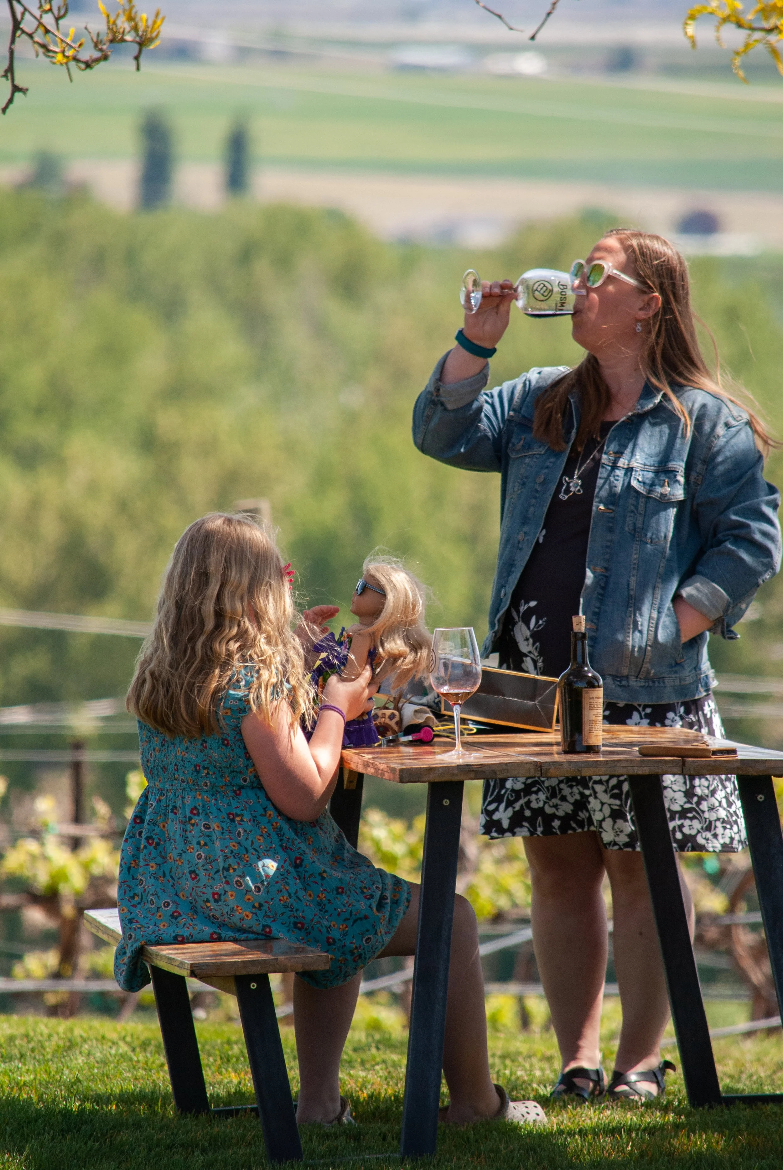 Two young girls with long hair, one with glasses, and an adult woman with glasses, all standing around a wooden outdoor table set in a green, rural landscape. The woman is drinking from a glass wine goblet, and the girl in the blue dress is holding a doll. The scene appears to be a casual outdoor gathering or picnic with wine and dolls, during daytime.