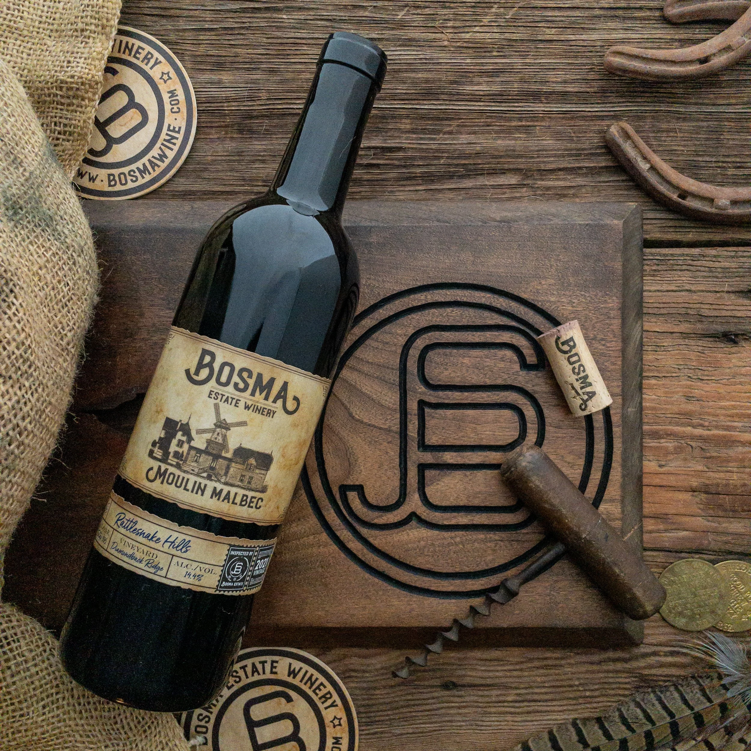 A wooden surface with a pair of sunglasses, animal hoof print shaped paper coaster, burlap sack, and a wine cork that reads 'Bosma Estate Winery' and 'Moulin Malbec'.