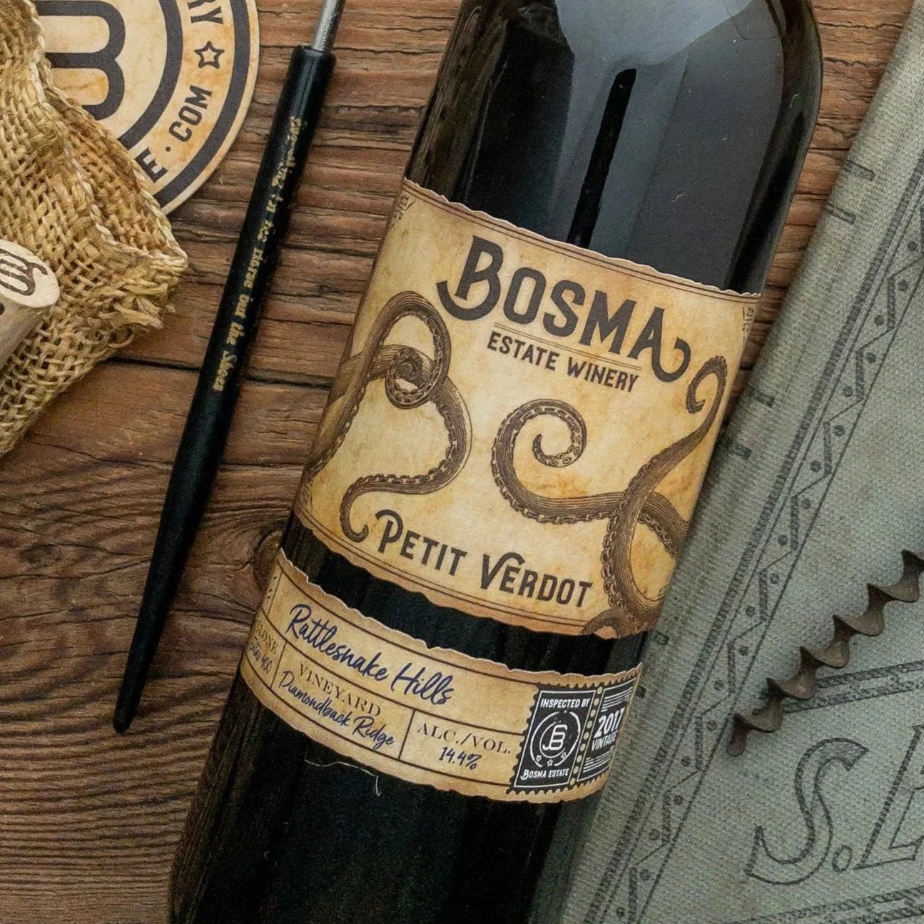A bottle of Bosma Estate Winery Petit Verdot wine on a rustic wooden surface, with a pen, a burlap sack, a coaster, and a cloth napkin nearby.
