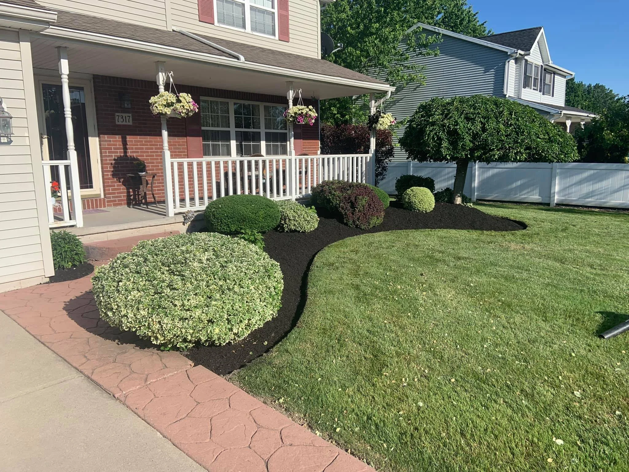 A neatly landscaped front yard with a garden bed containing various bushes and a small tree, bordered by a curved strip of dark mulch, with a porch and a white fence in the background.  DB Property Maintenance.