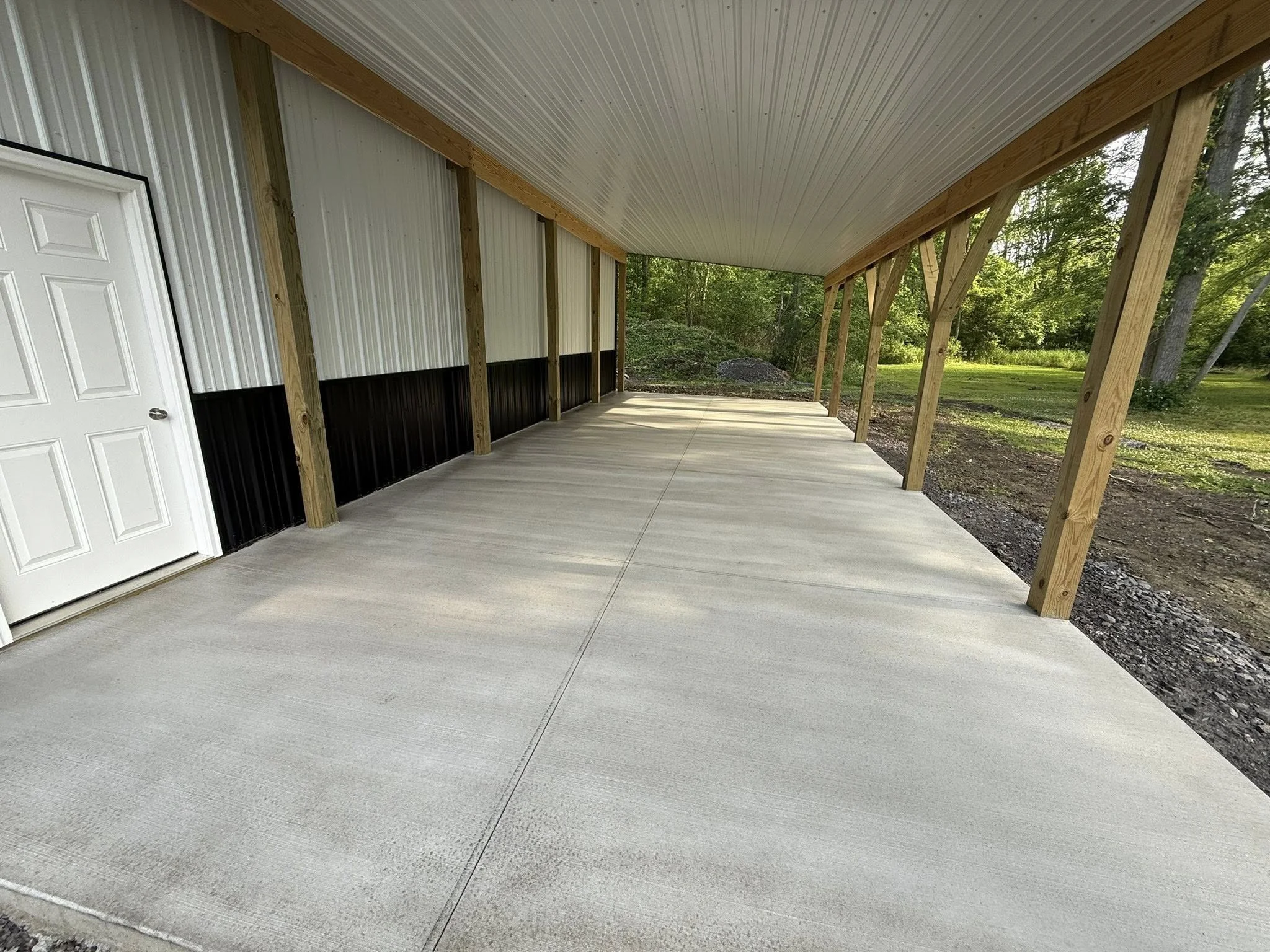 A covered outdoor patio with a concrete floor, supported by wooden beams, attached to a building with white and black siding, surrounded by trees and greenery. DB Property Maintenance.