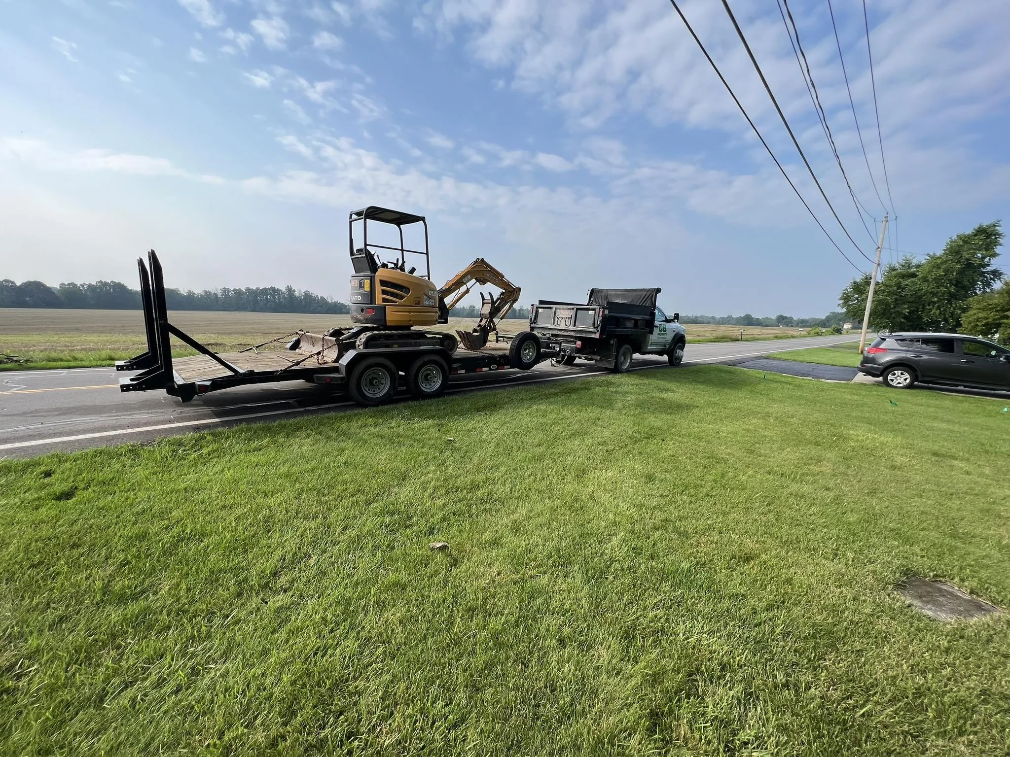A trailer carrying a small excavator and other construction equipment is parked on the side of a rural road. A silver truck is attached to the trailer, and a black SUV is parked nearby.  DB Property Maintenance.