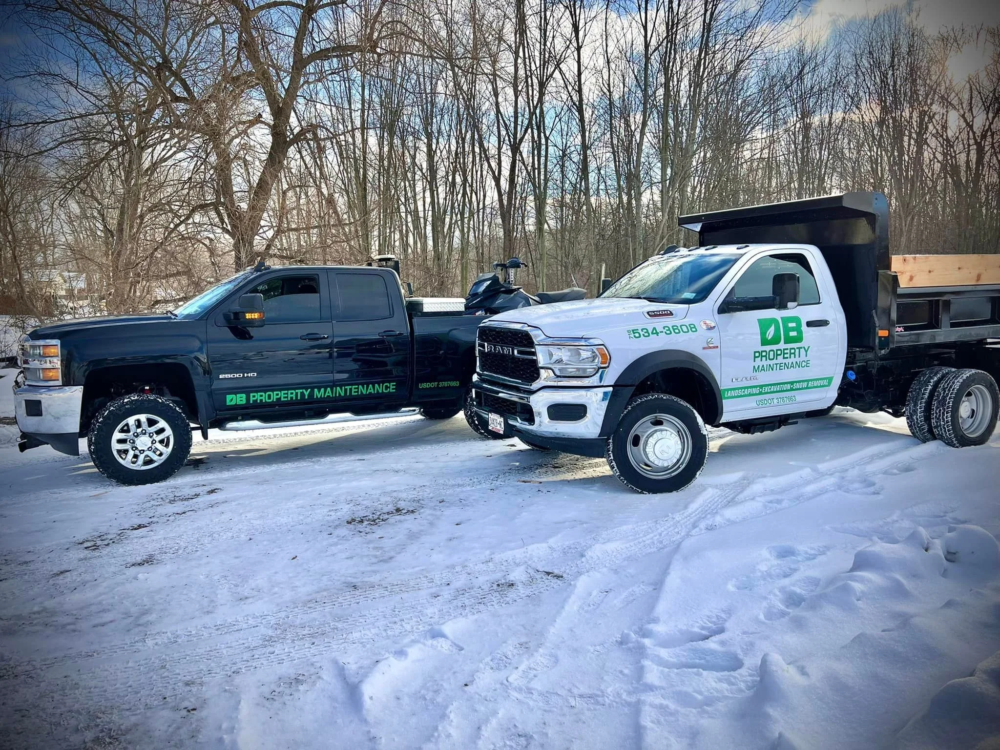 Two service trucks, one black and one white, parked on snow-covered ground with leafless trees in the background. The trucks have green and white branding for DB Property Maintenance.