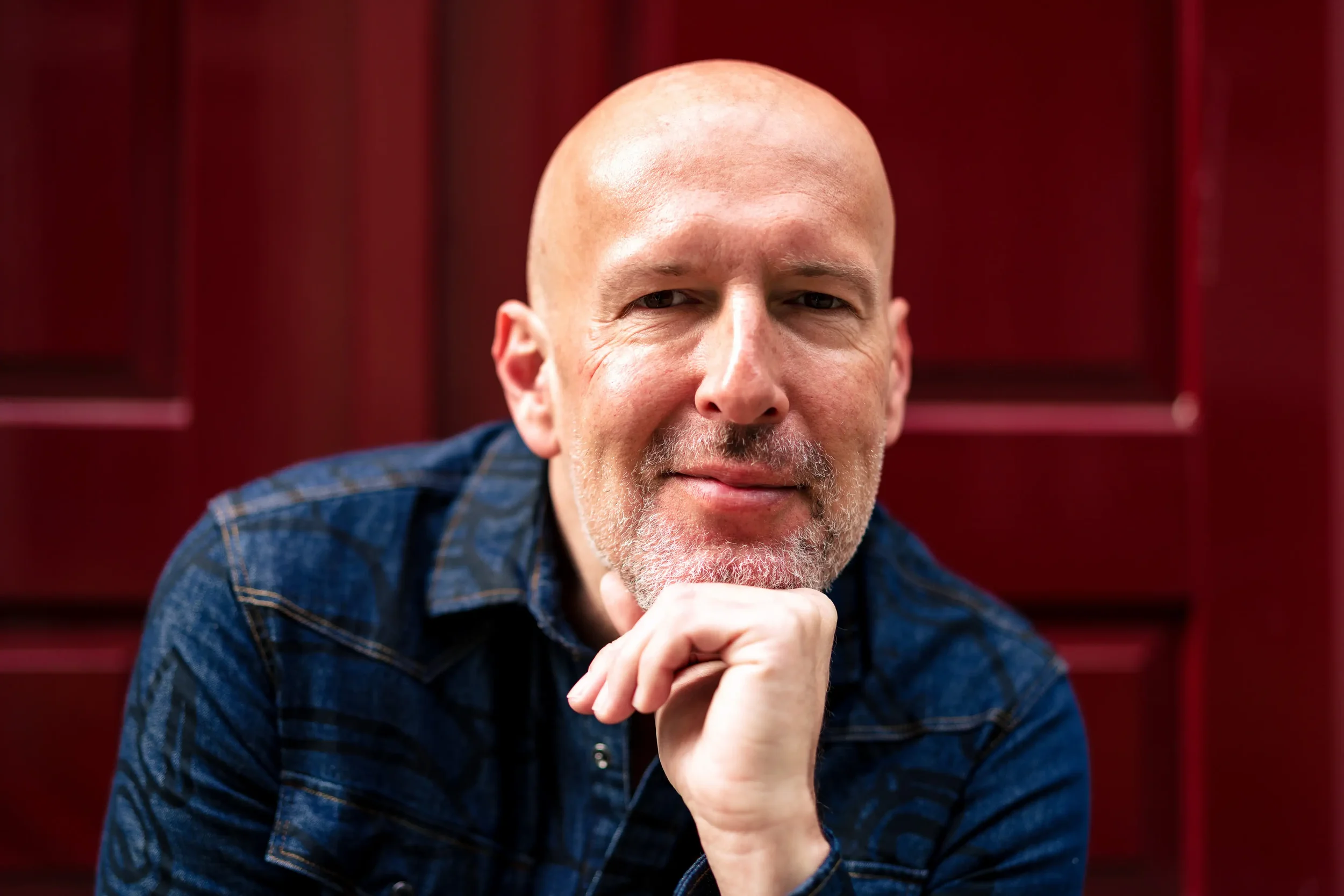 Close-up van een man met een kale kop en een grijze baard, poseert met zijn hand onder zijn kin voor een rode achtergrond.