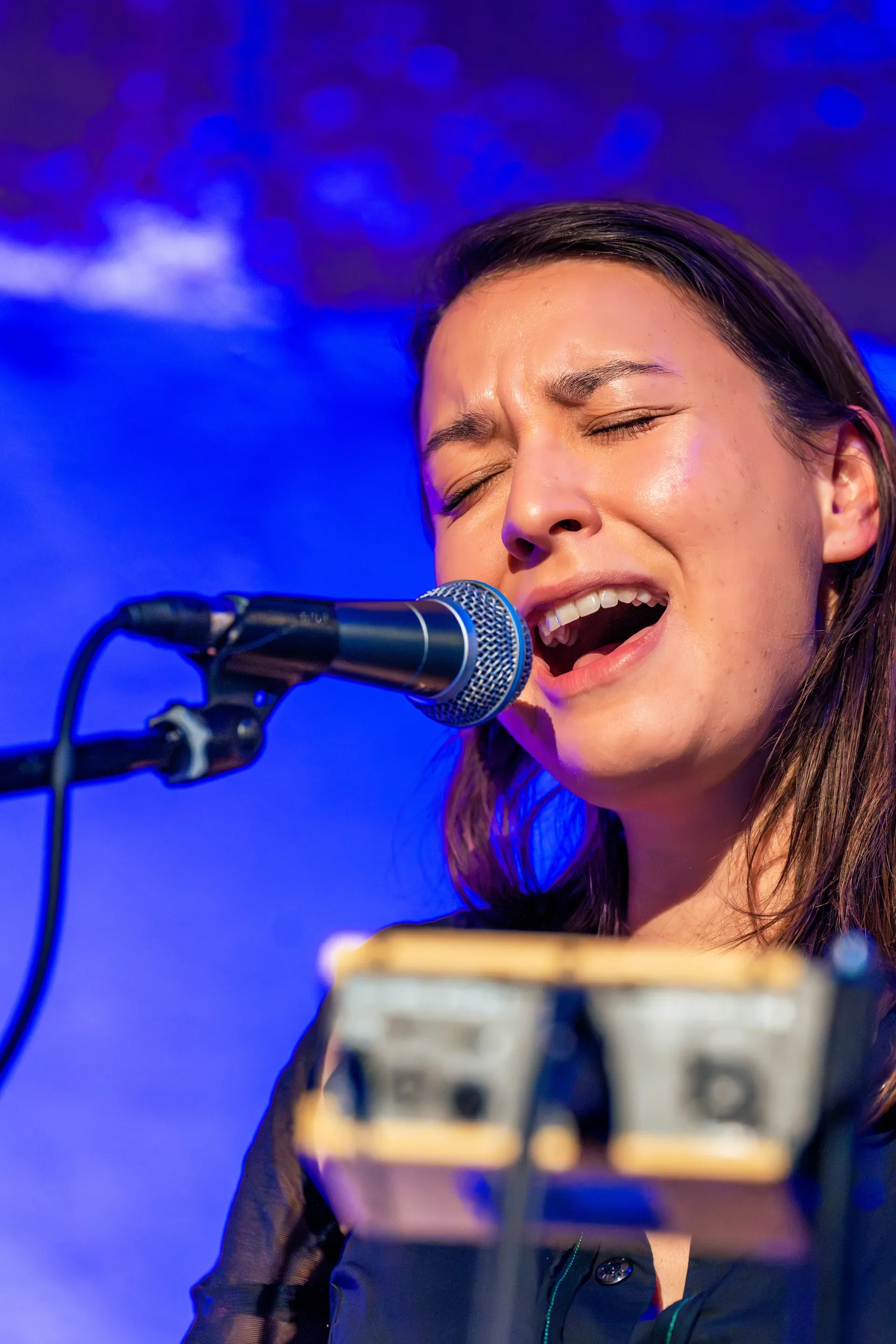 Een jonge vrouw zingt met haar ogen dicht achter een microfoon op een blauwe achtergrond.