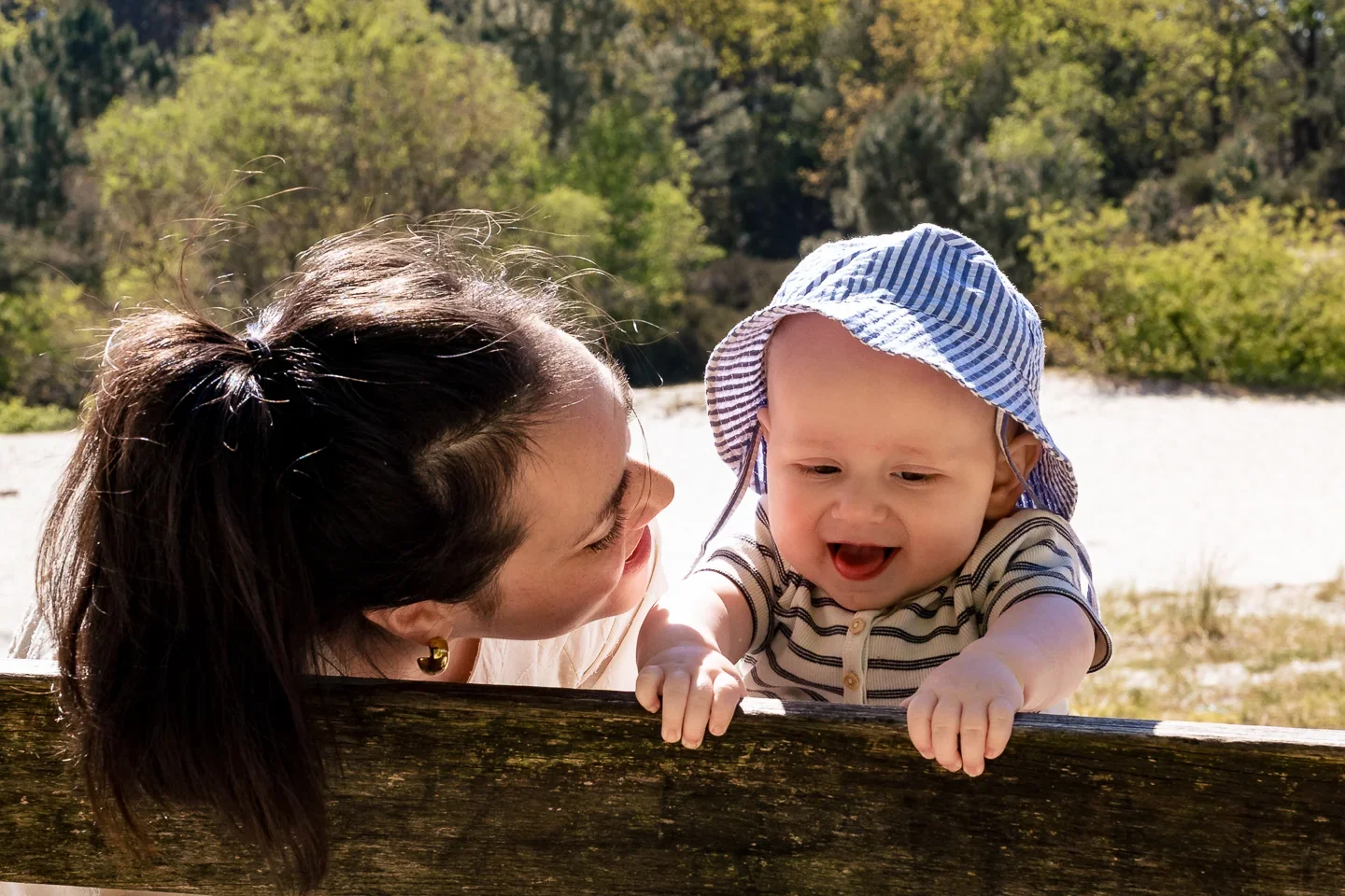 Een vrouw en een baby lachen en spelen buiten in een natuurlijke omgeving met bomen en water op de achtergrond. De baby draagt een blauwe hoed en een gestreept shirt.