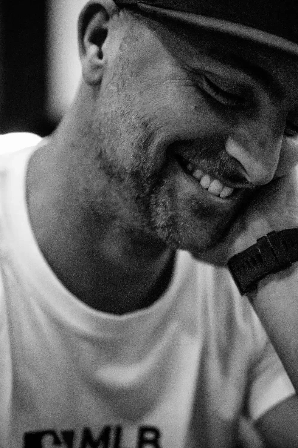 Close-up zwart-wit foto van lachende man met baard, schattig zeggend, terwijl hij zijn hoofd op zijn hand rust.