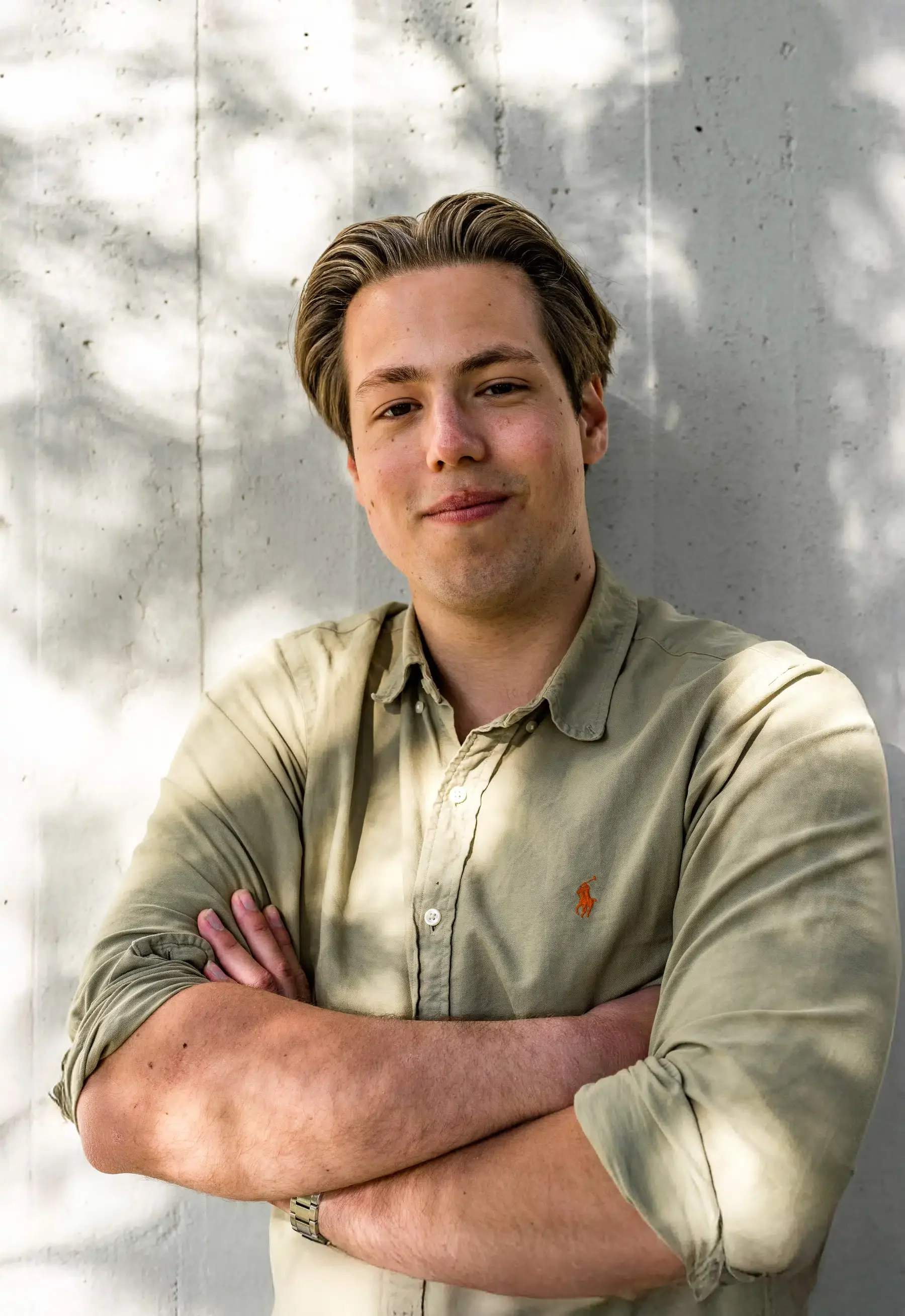 Portret van een jonge man met gevlochten haar, staand voor een witte muur met schaduw van bladeren.