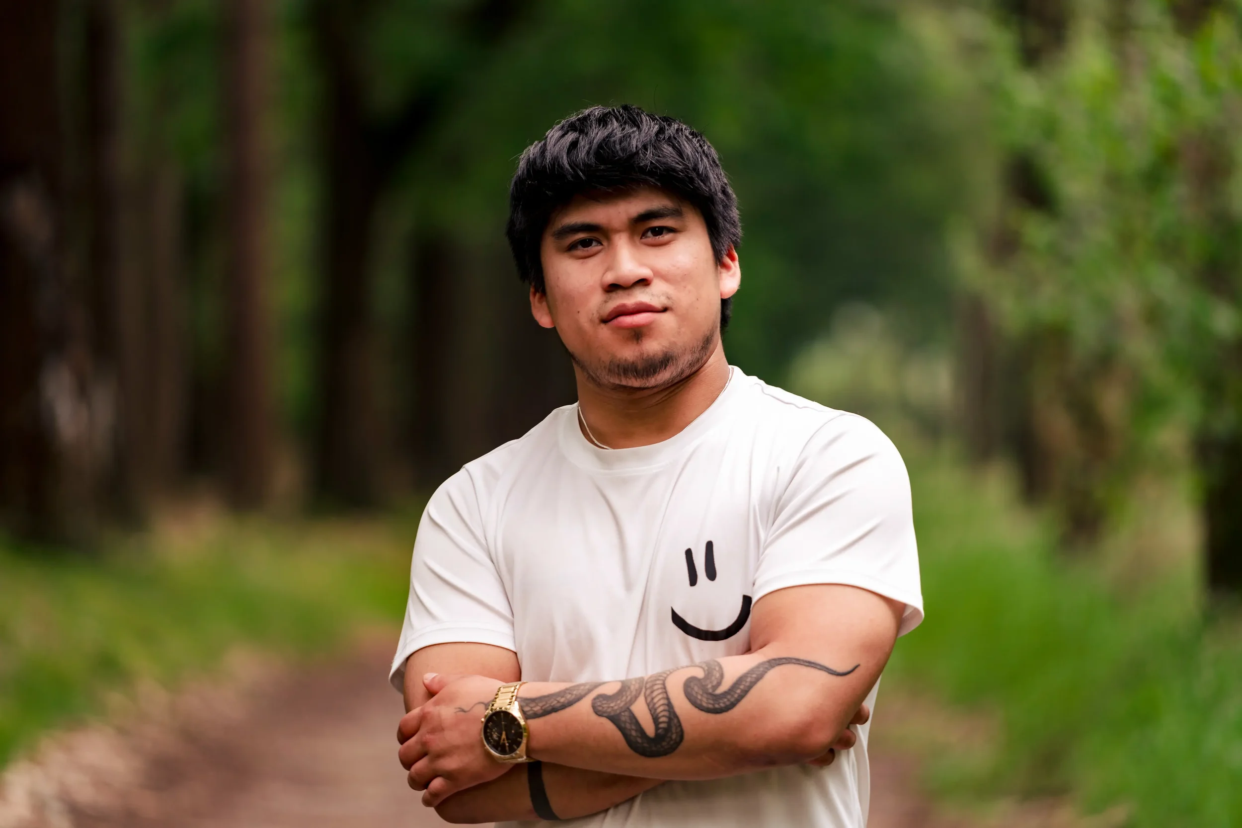 Jonge man met zwart haar en een lichte huid, staand met gekruiste armen in een bos, draagt een witte t-shirt met een zwarte smiley op de borst en een gouden horloge, met een serpent-tattoo op zijn linker onderarm.