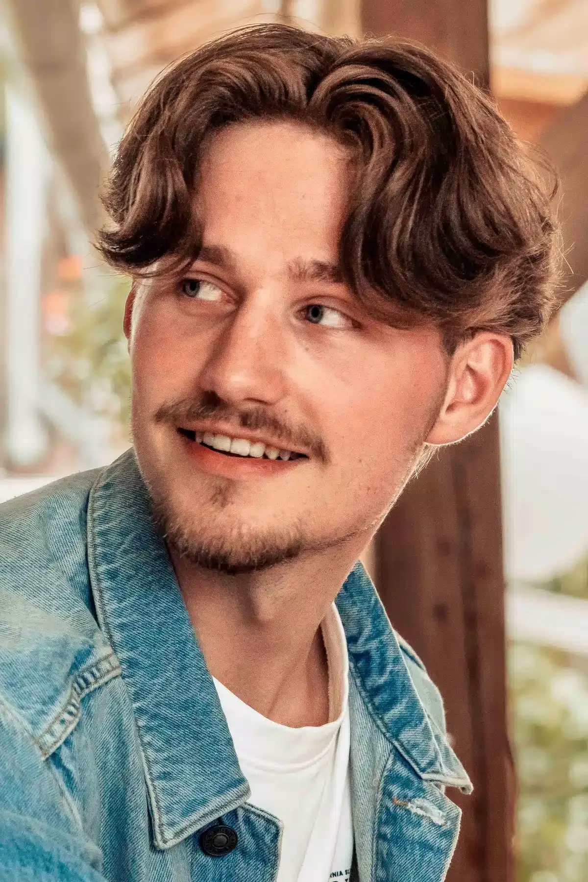 Een jonge man met bruin, golvend haar, een lichte huidskleur en een lichte baard, draagt een blauwe spijkerjack en een witte t-shirt. Hij kijkt naar links, glimlacht en zit voor een raam met een tuin op de achtergrond.