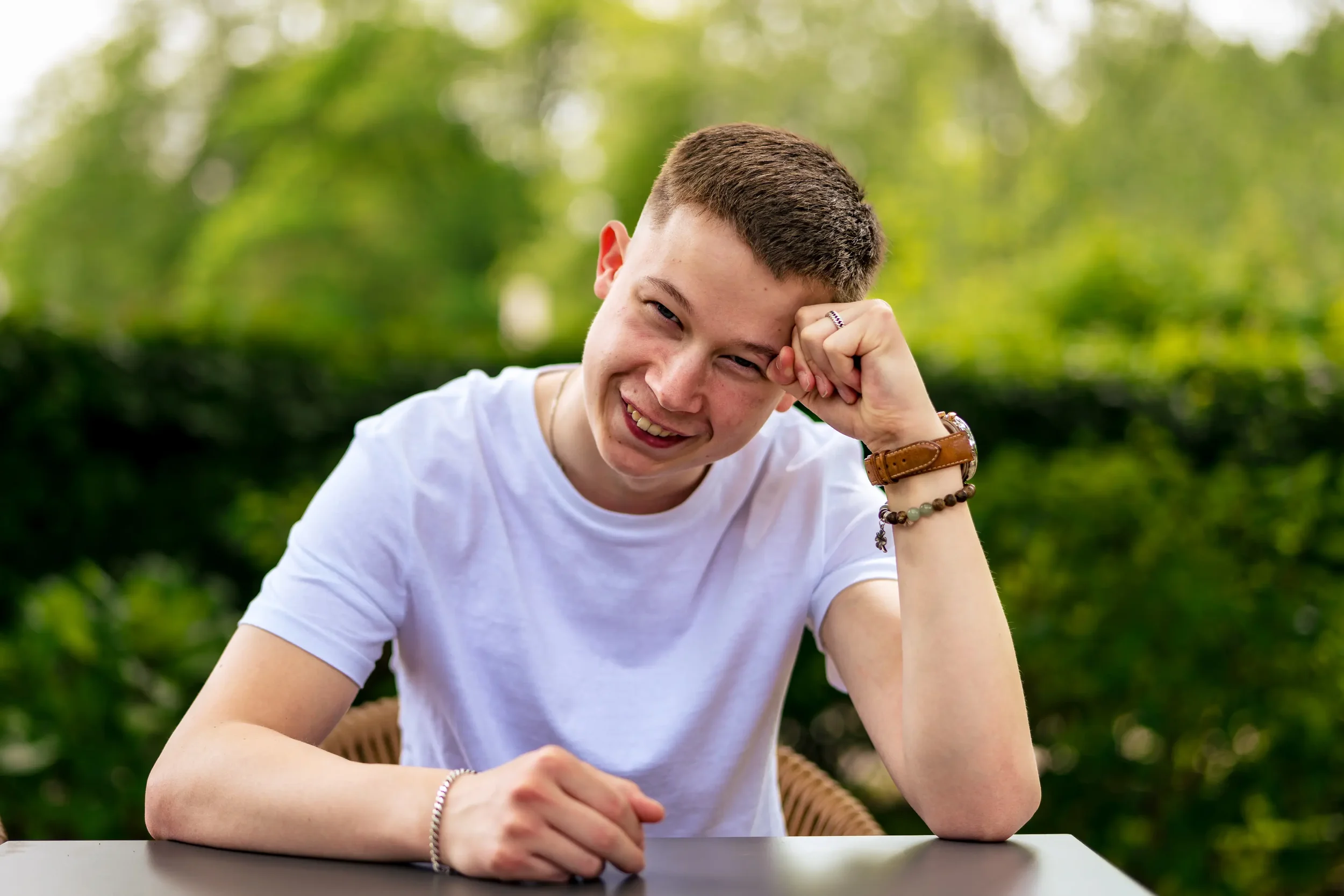 Een glimlachende jonge man met kort haar, zittend aan een tafel in een groene, buitenomgeving, met gekleed in een witte T-shirt en accessoires zoals horloges en armbanden.