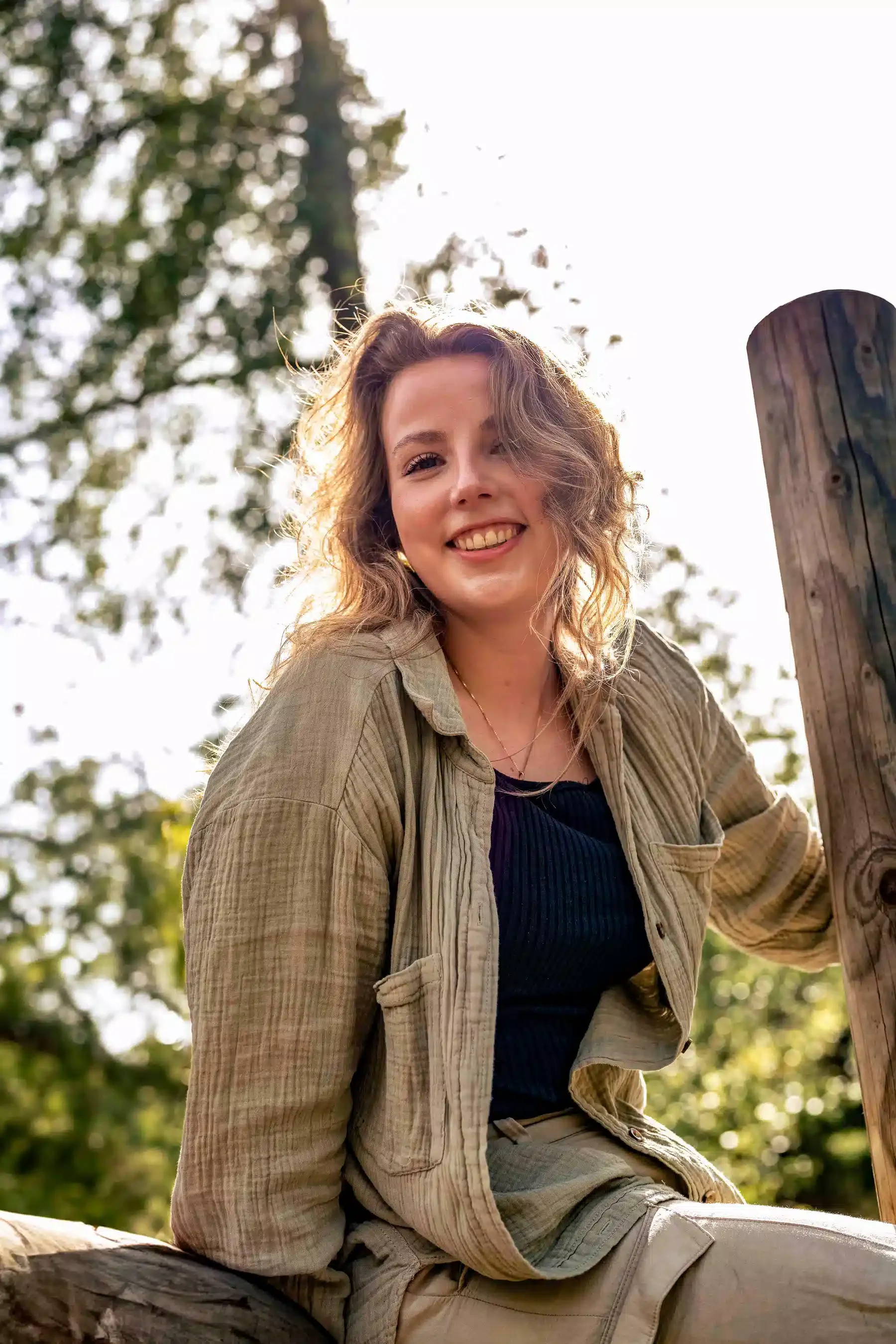Een lachende jonge vrouw met golvend bruin haar zit buiten op een boomstam, met een houten paal naast haar en een bos met bomen op de achtergrond, onder een heldere hemel.