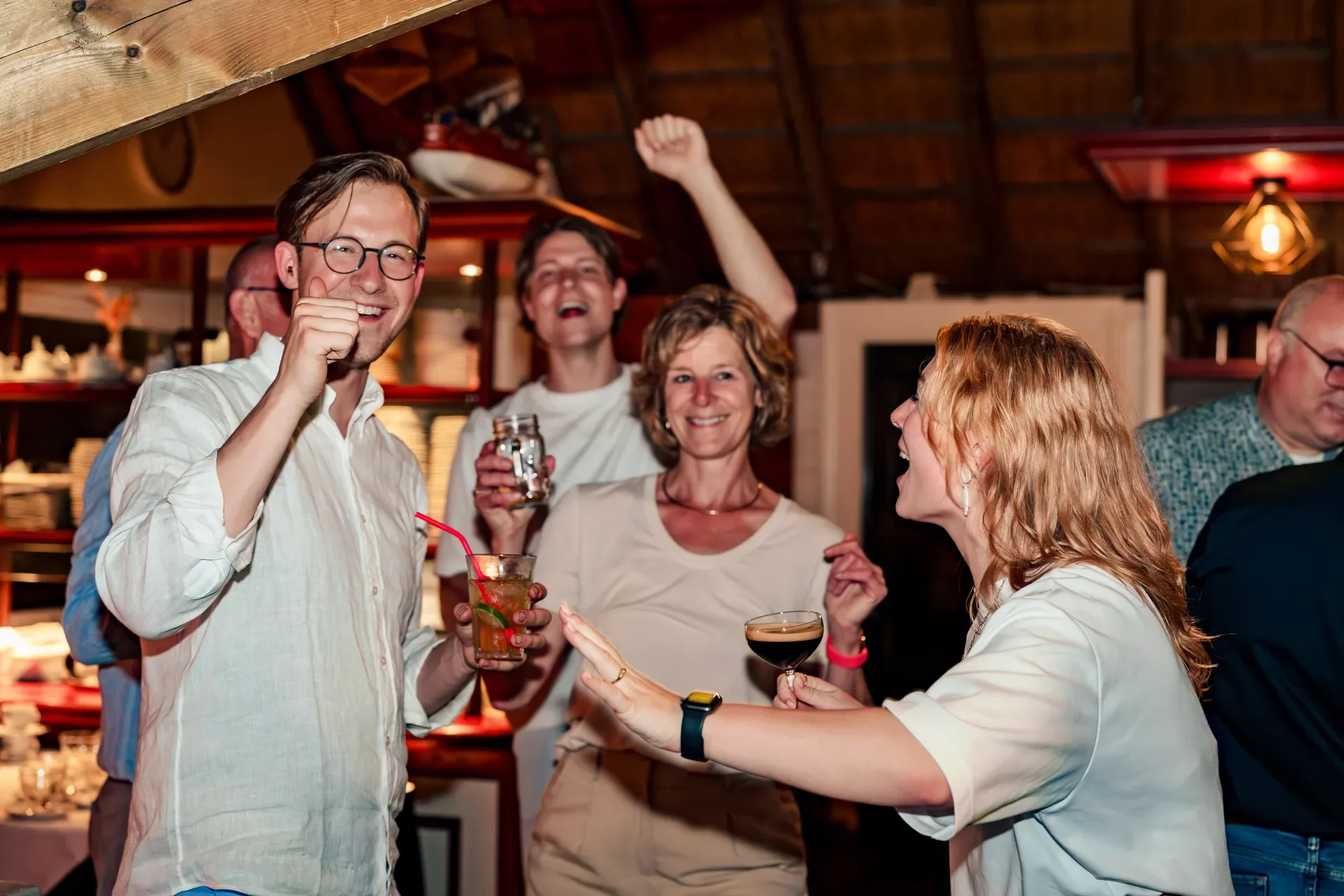 Vijf mensen die genieten van een feestje in een gezellige, warme sfeer, met glazen drank in hun handen en lachen.