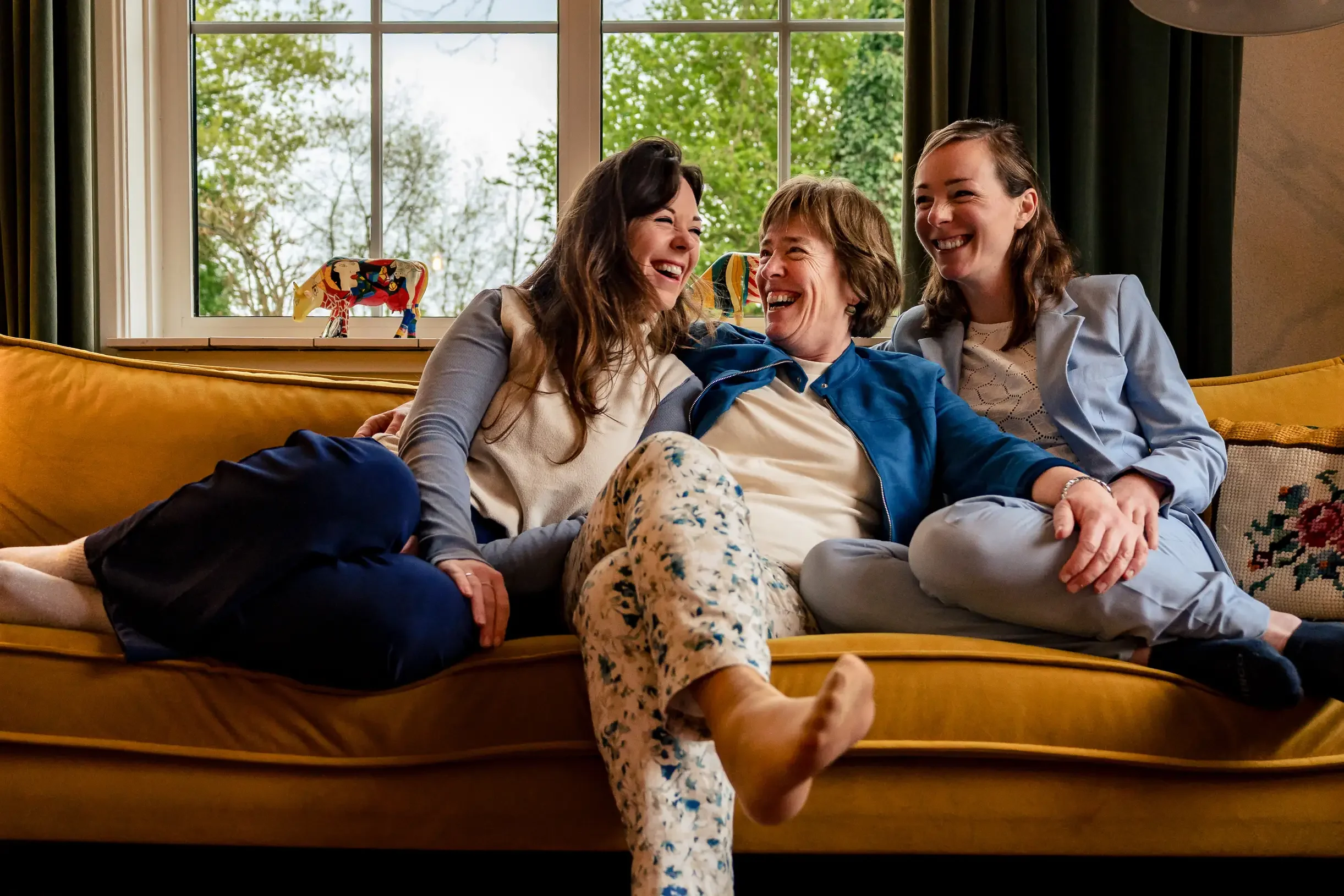 Drie vrouwen zitten samen op een gele bank, lachen en hebben plezier in een woon- of woonkamer met een groot raam en uitzicht op bomen.