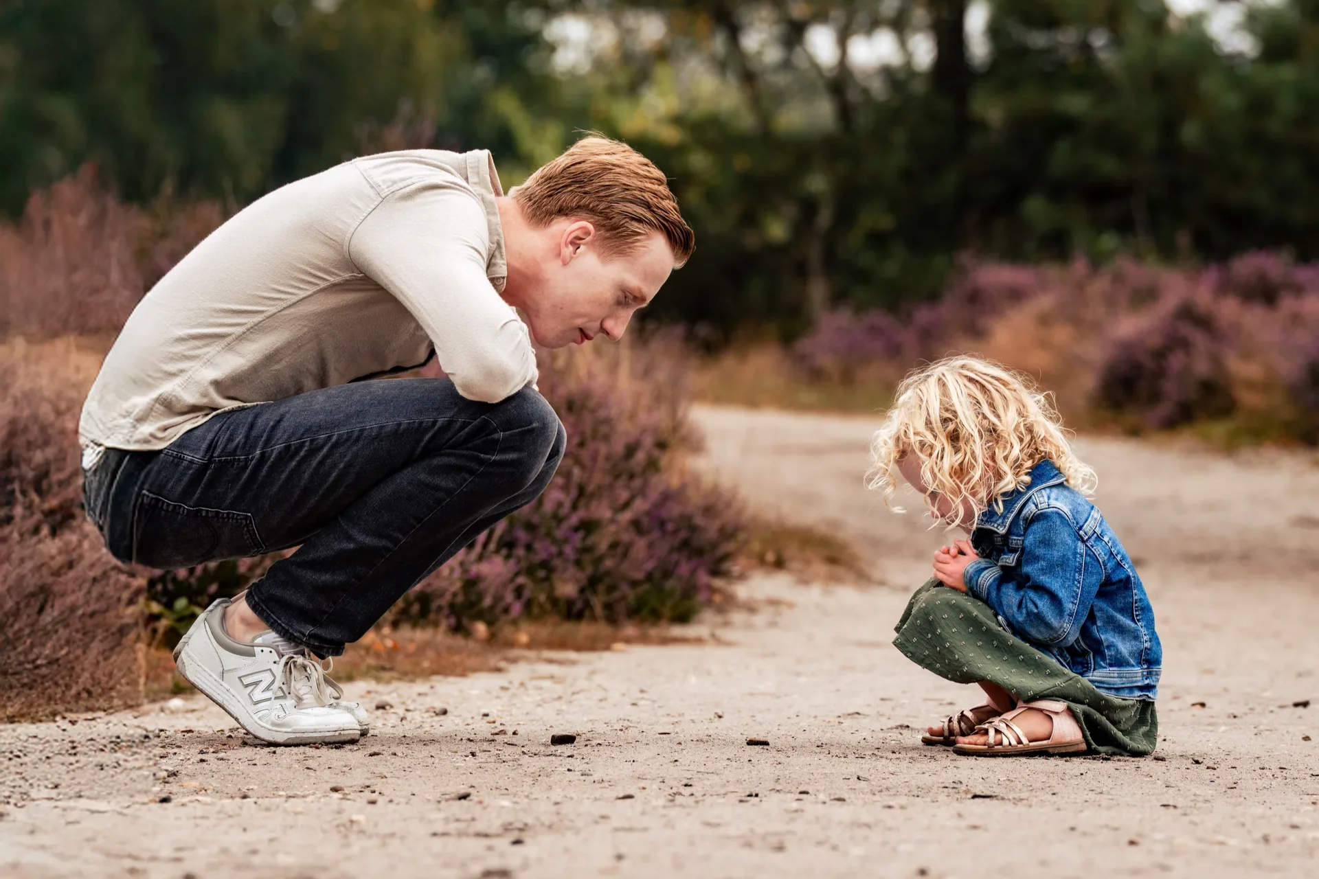 Man knielt voor een meisje dat op de grond zit, in een natuurlijke omgeving met struiken en bomen, beide kijken naar kleine voorwerpen op de grond.