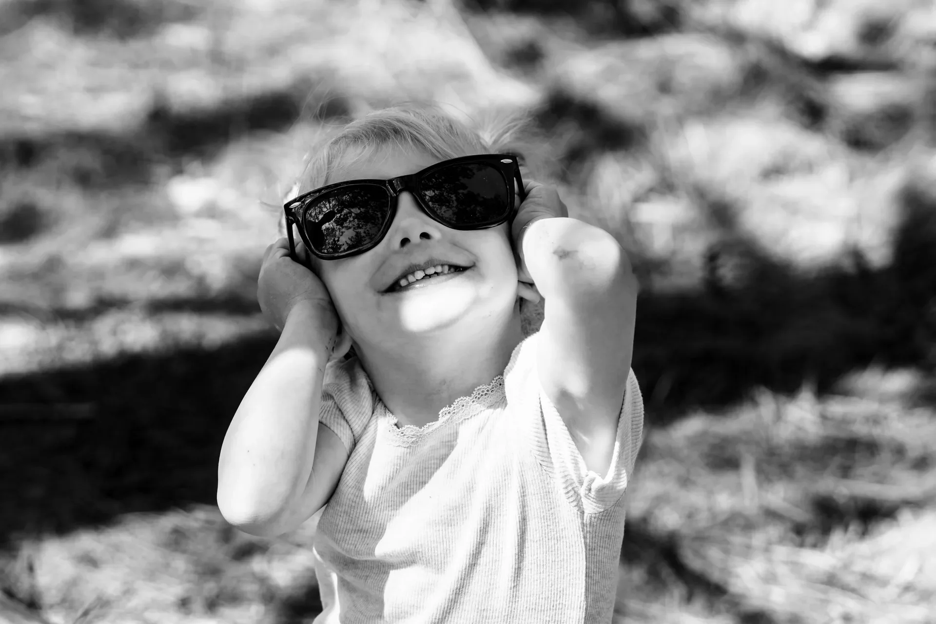 Een glimlachende kind met blonde haar en grote zonnebril, zittend buiten op een zonnige dag.