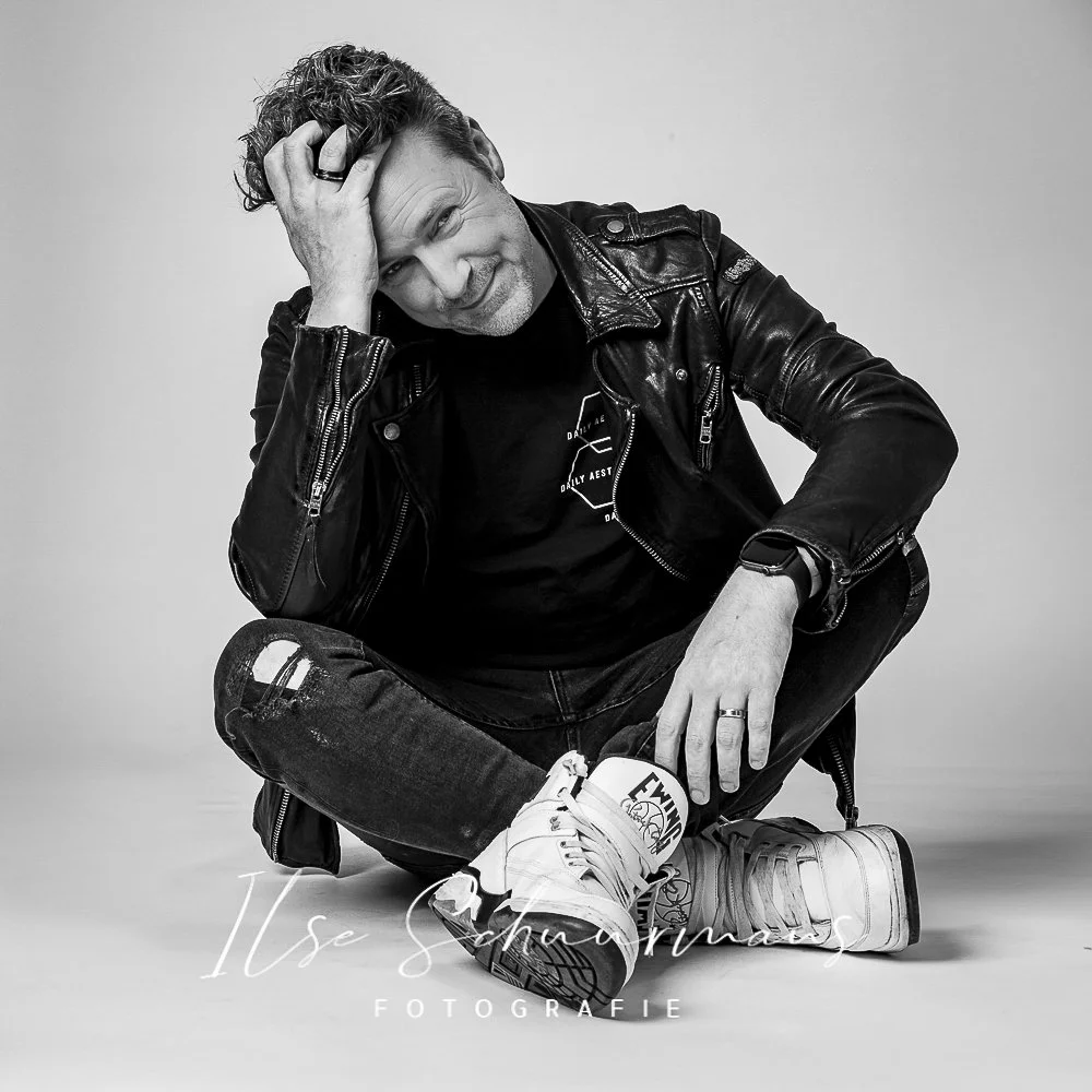 Zittende man in een leren jas, met een hand door zijn haar, glimlachend, op een witte achtergrond, zwarte en witte foto.