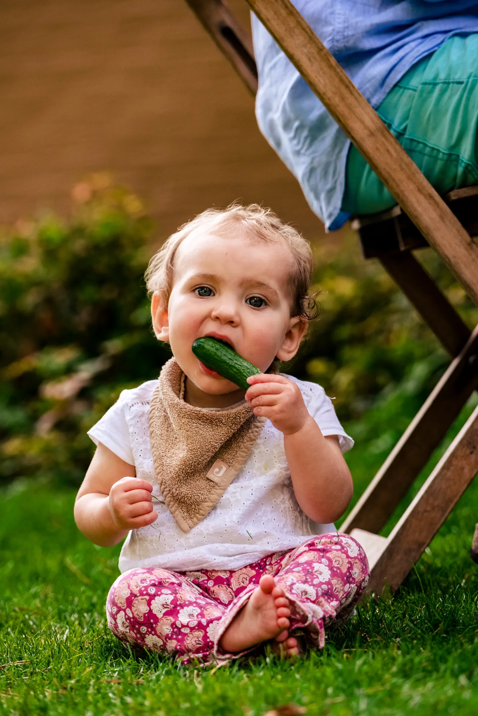 Een jonge kind zit op het gras en bijt in een komkommer.