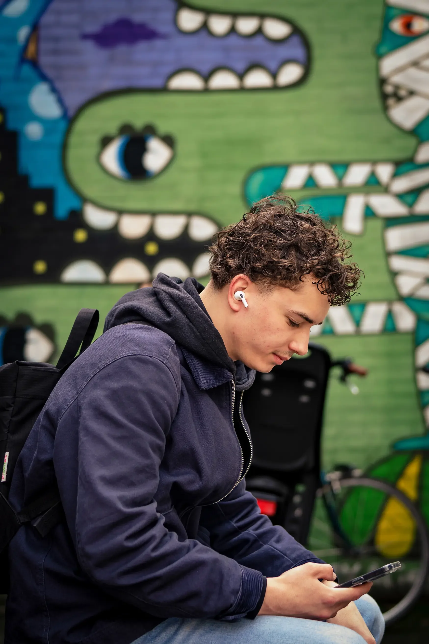 Een jonge man met krullend haar zit buiten, kijkt op zijn telefoon en draagt oordopjes, met een kleurrijke muurschildering van een drakenvuur in de achtergrond.