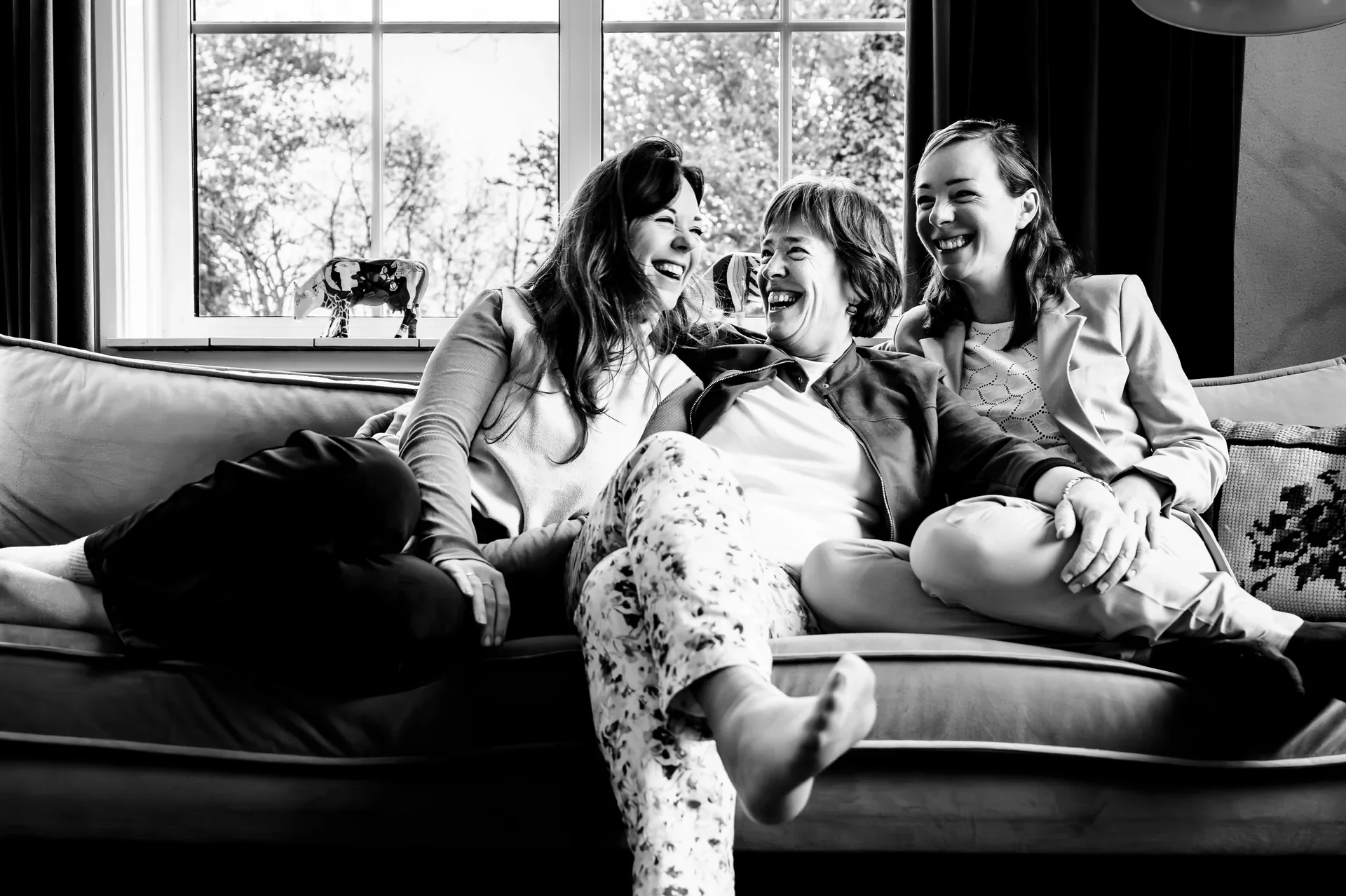 Drie vrouwen die lachend op een bank zitten in een woonkamer met grote ramen en een uitzicht op bomen buiten.