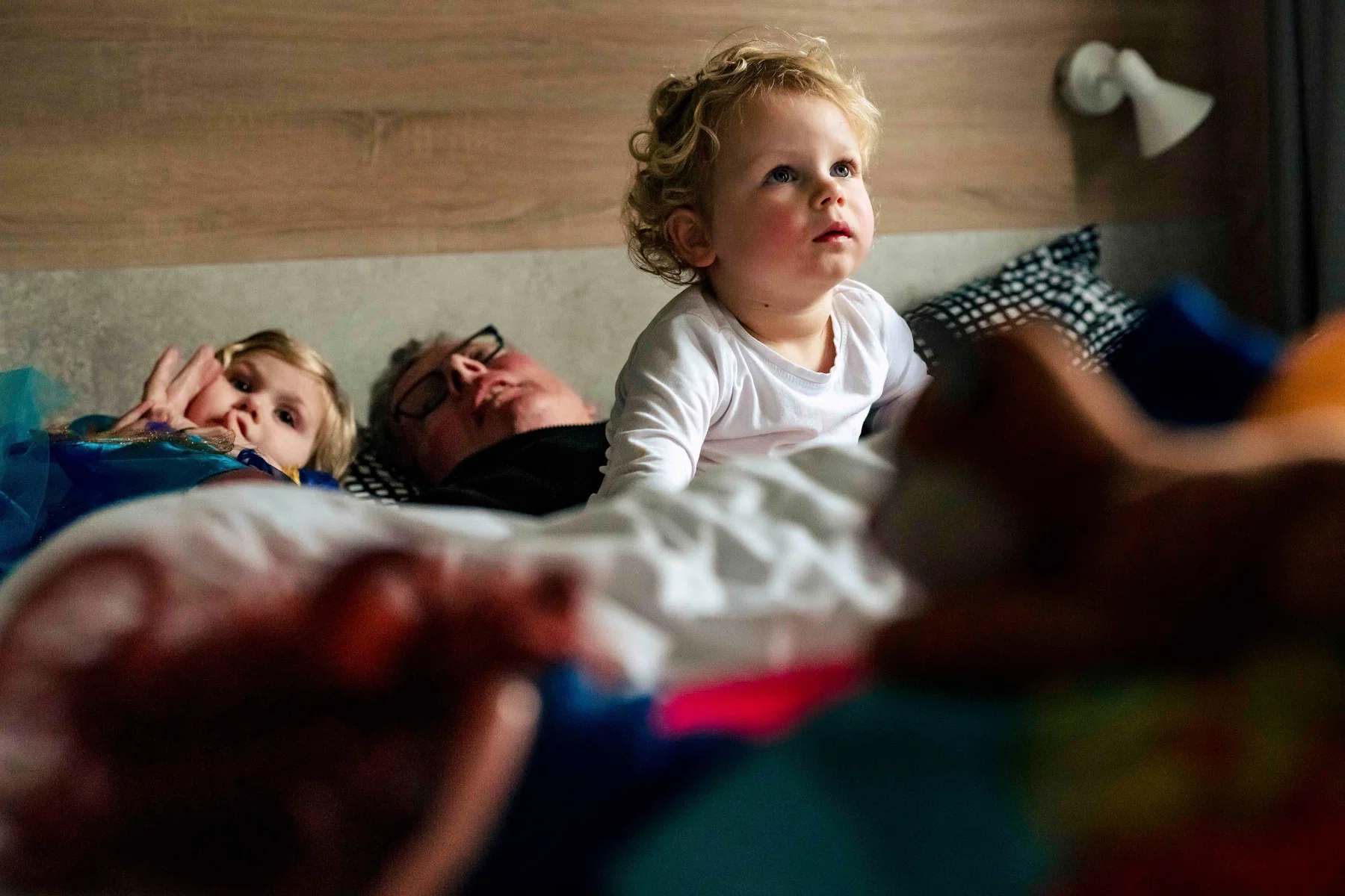 Twee jonge kinderen en een man liggen op een bed, de kinderen kijken geïnteresseerd of verwonderd, de man ligt met zijn ogen dicht.