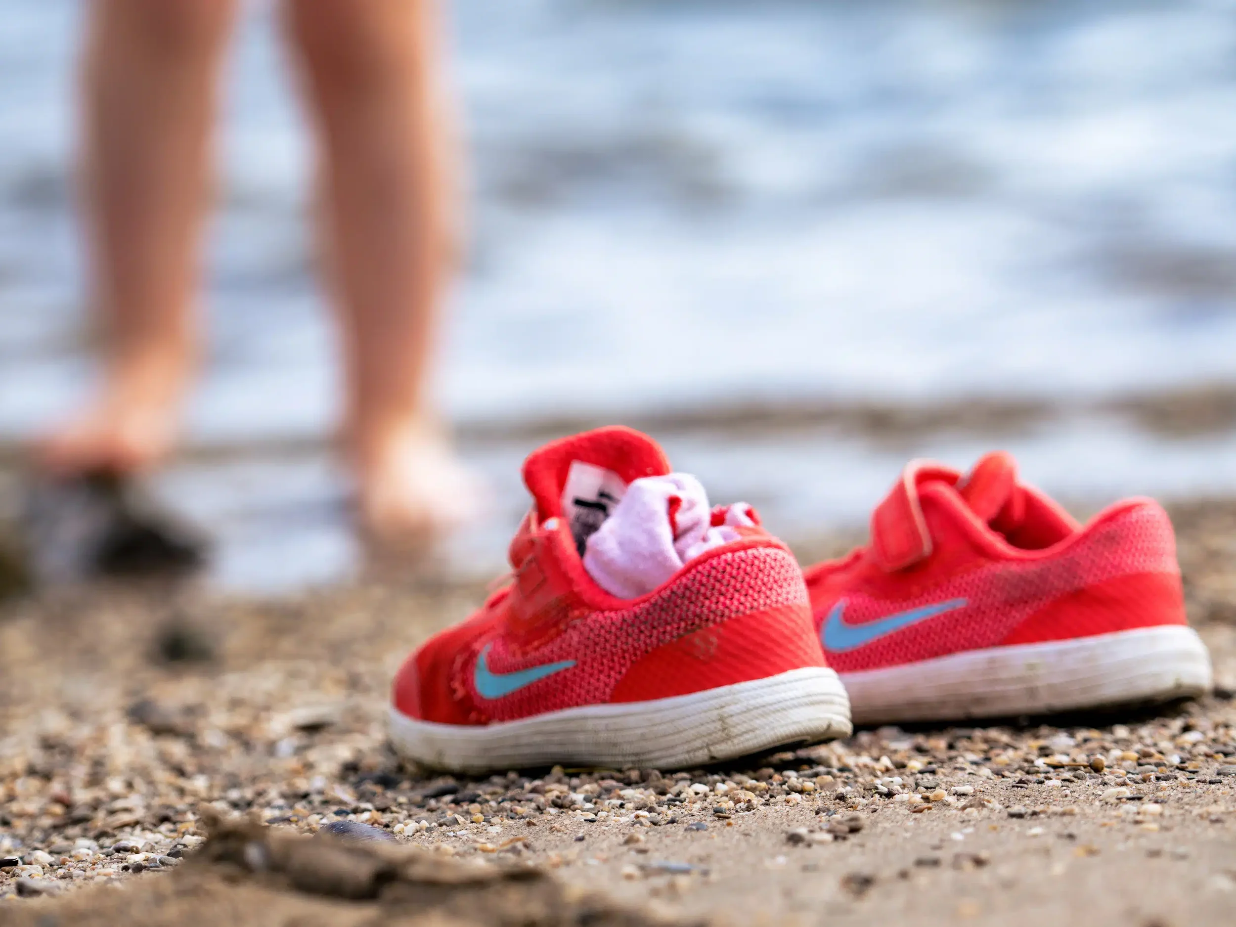 Een paar roze sportschoenen en een wit en roze sok liggen op het zand op het strand, met water en de benen van een kind in de achtergrond.