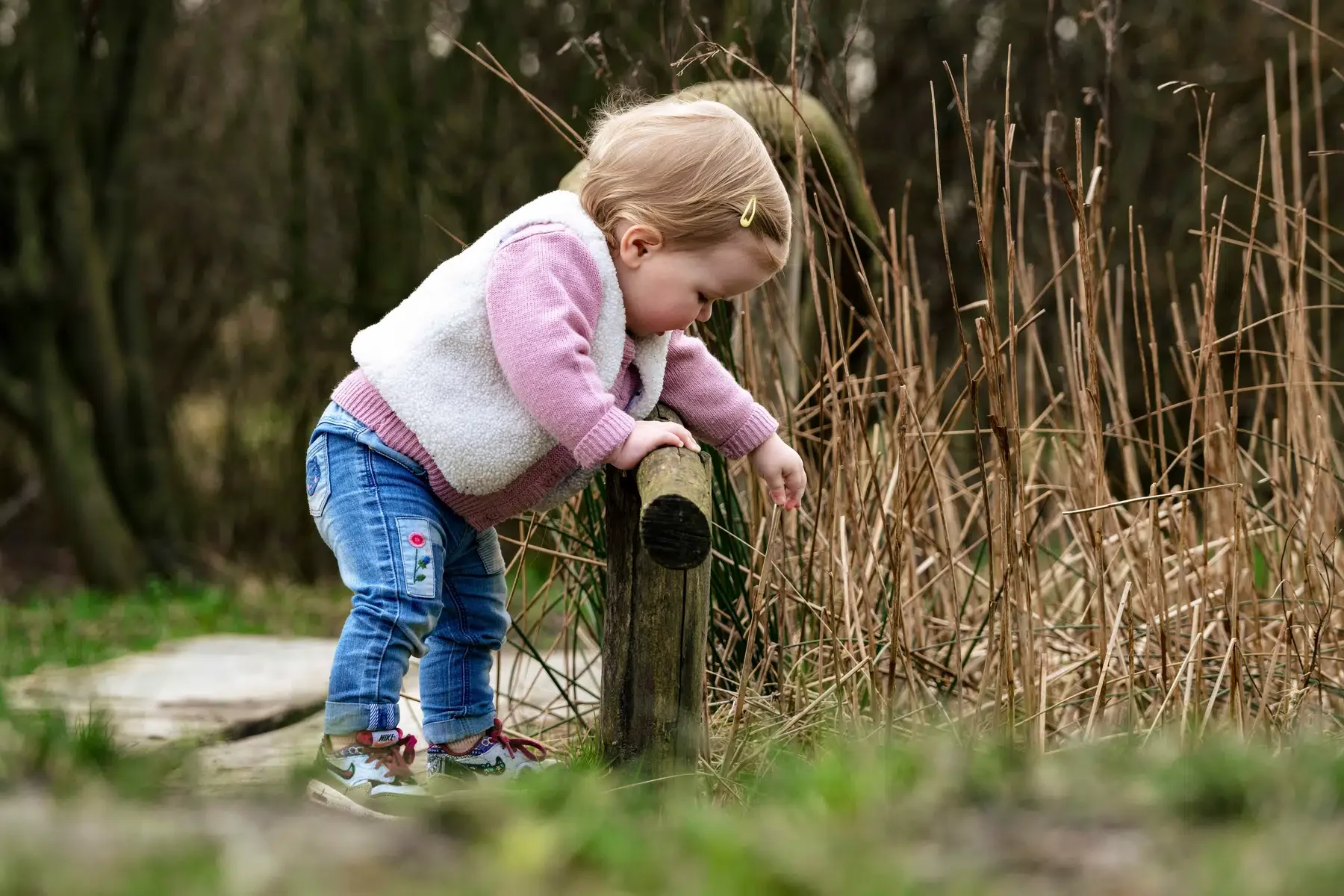 een jonge meisje dat op een bospad staat en een houten paal vasthoudt, omringd door gras en dorre planten in een natuurlijk buitengebied
