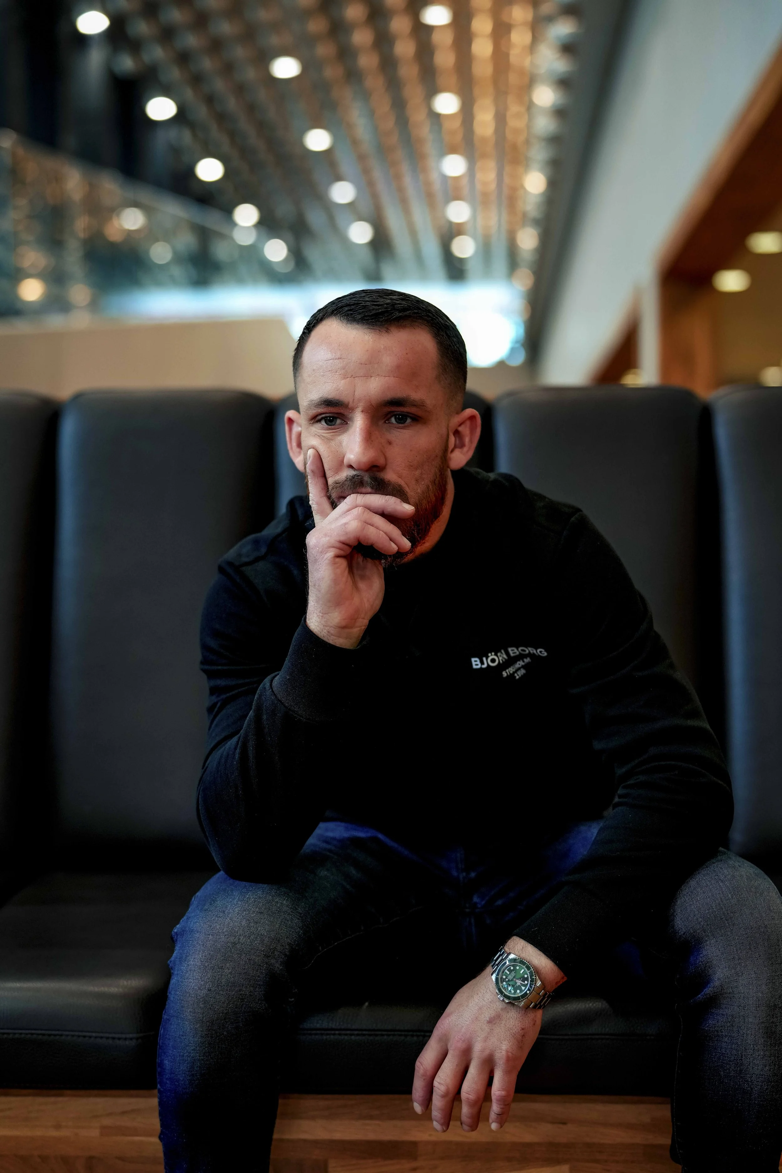 Man zittend op een bank in een moderne interieur met plafondverlichting, rustgevende uitstraling, denkende houding, zwarte jas en horloge.