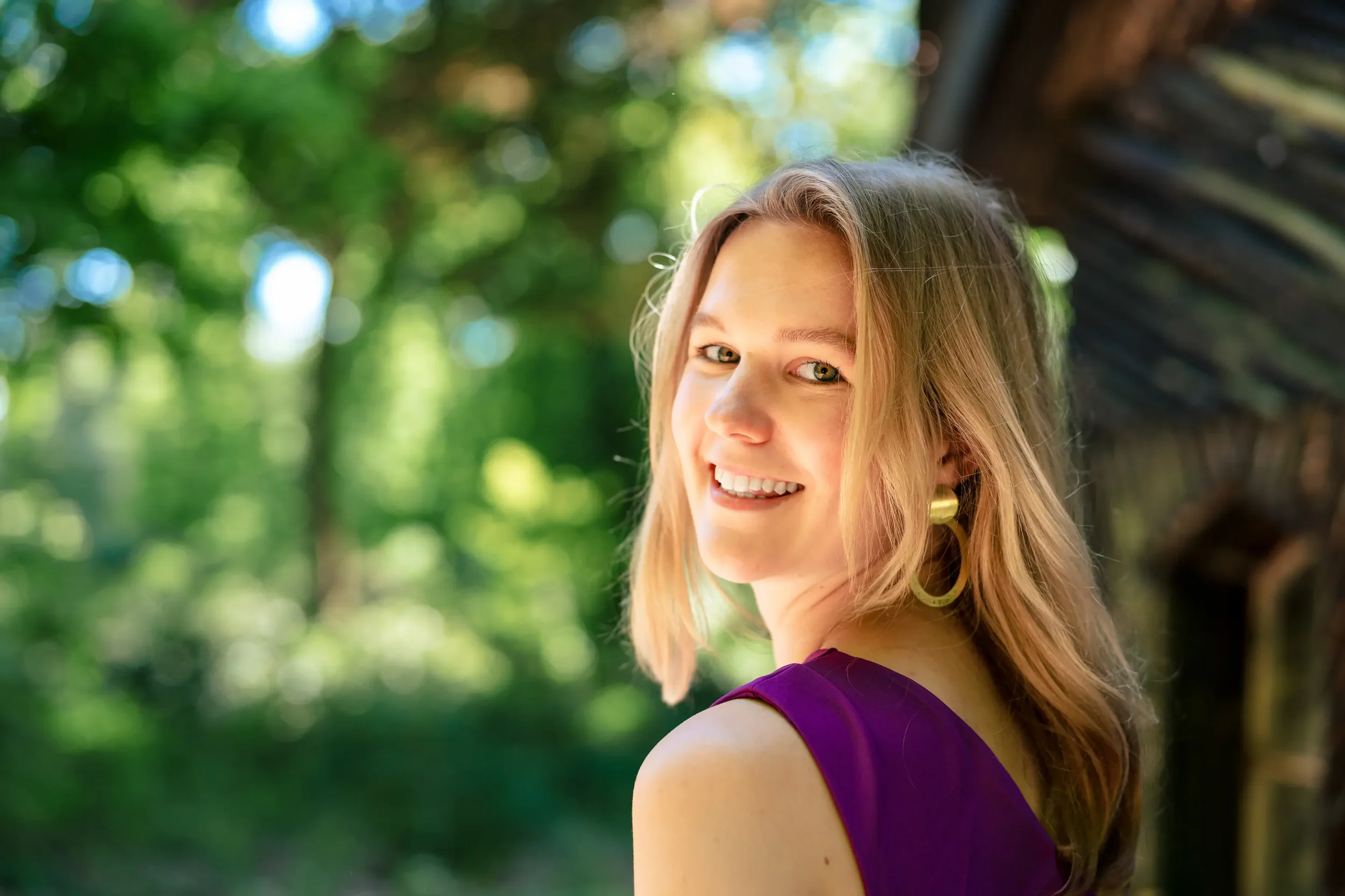 Een jonge vrouw met lichtbruin haar die glimlacht, in een groen bos, gekleed in een paarse jurk en grote oorbellen.